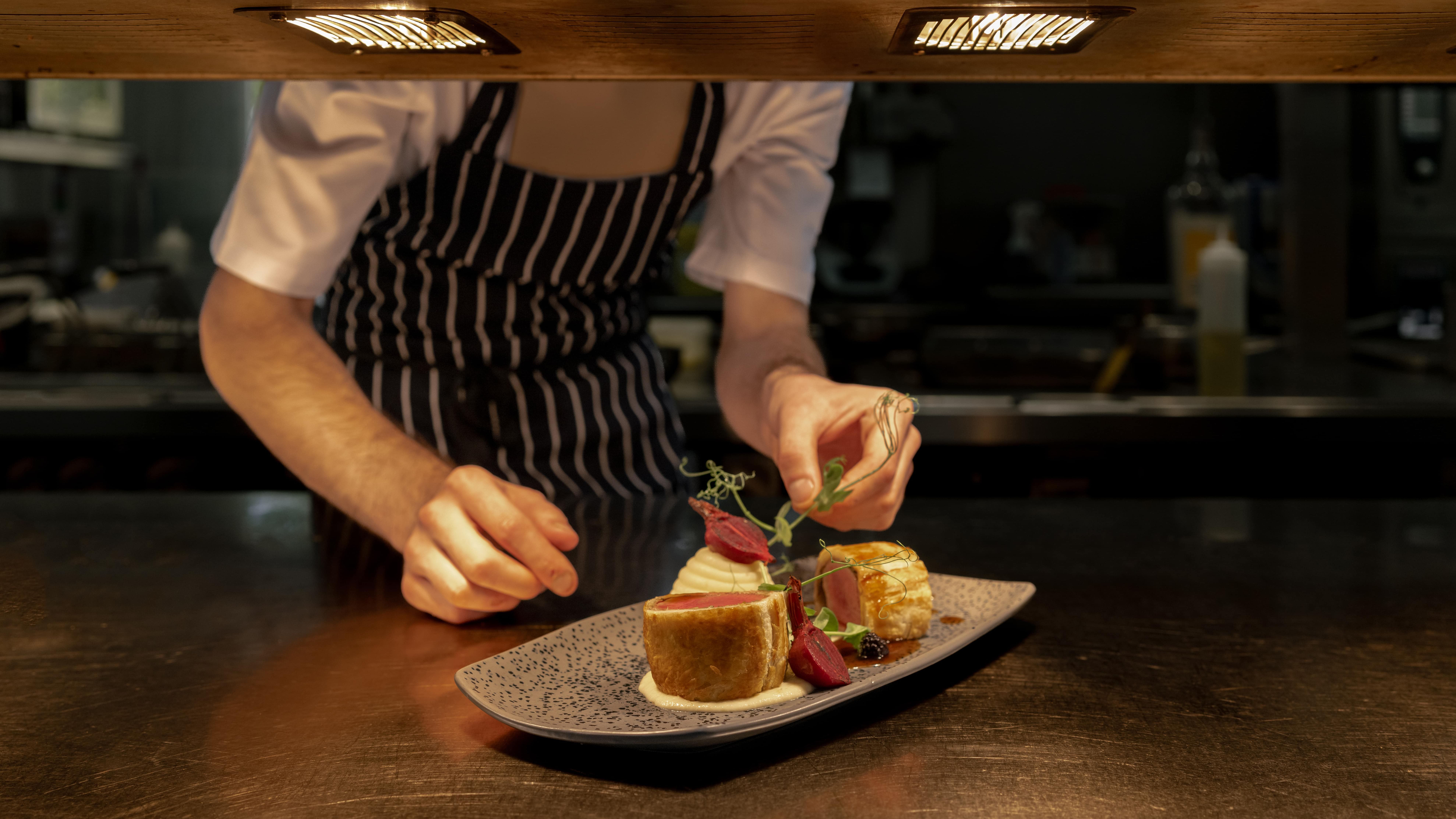 Chef in striped apron carefully garnishing a plated gourmet dish under kitchen lights