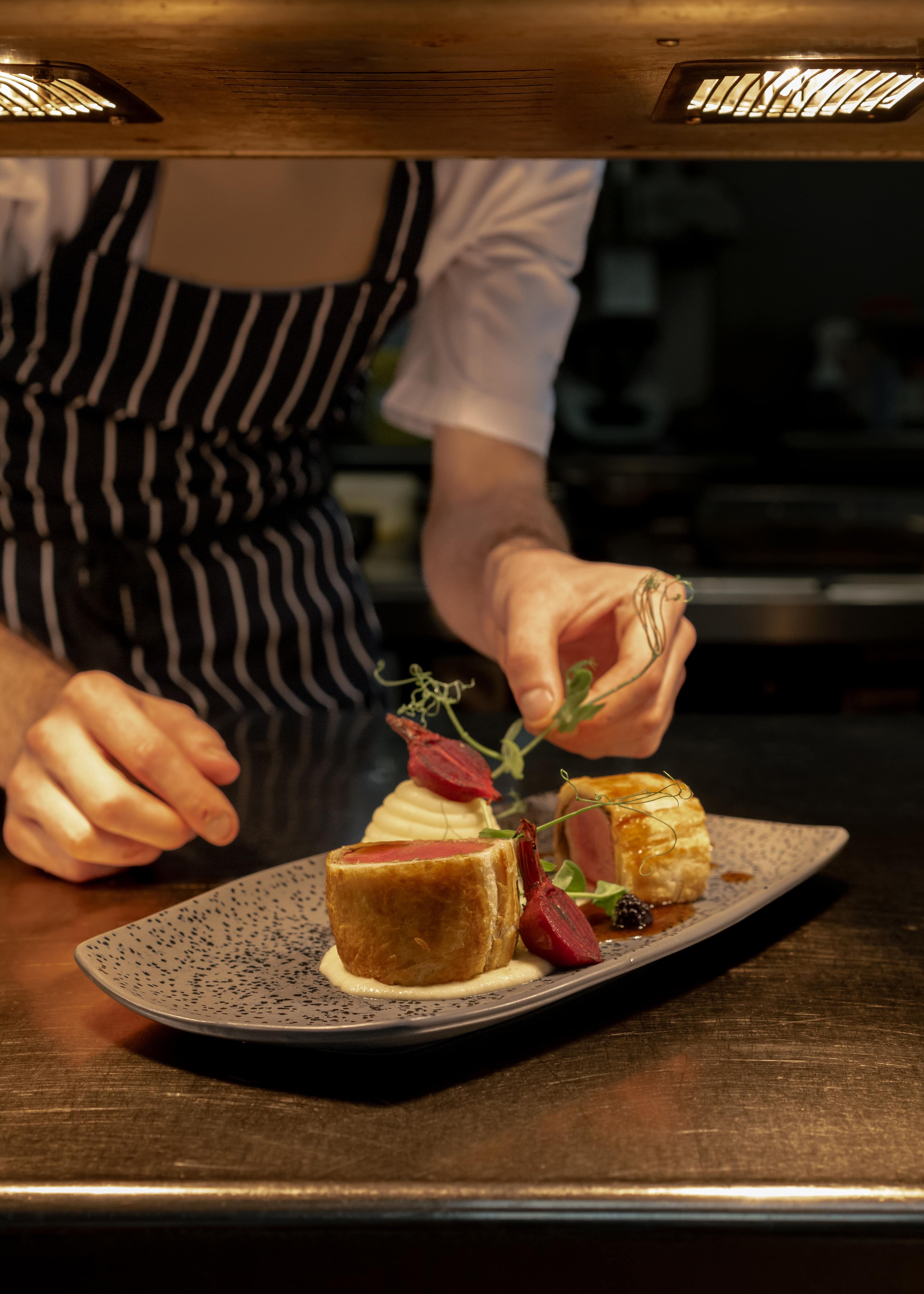 Chef in striped apron carefully garnishing a plated gourmet dish under kitchen lights