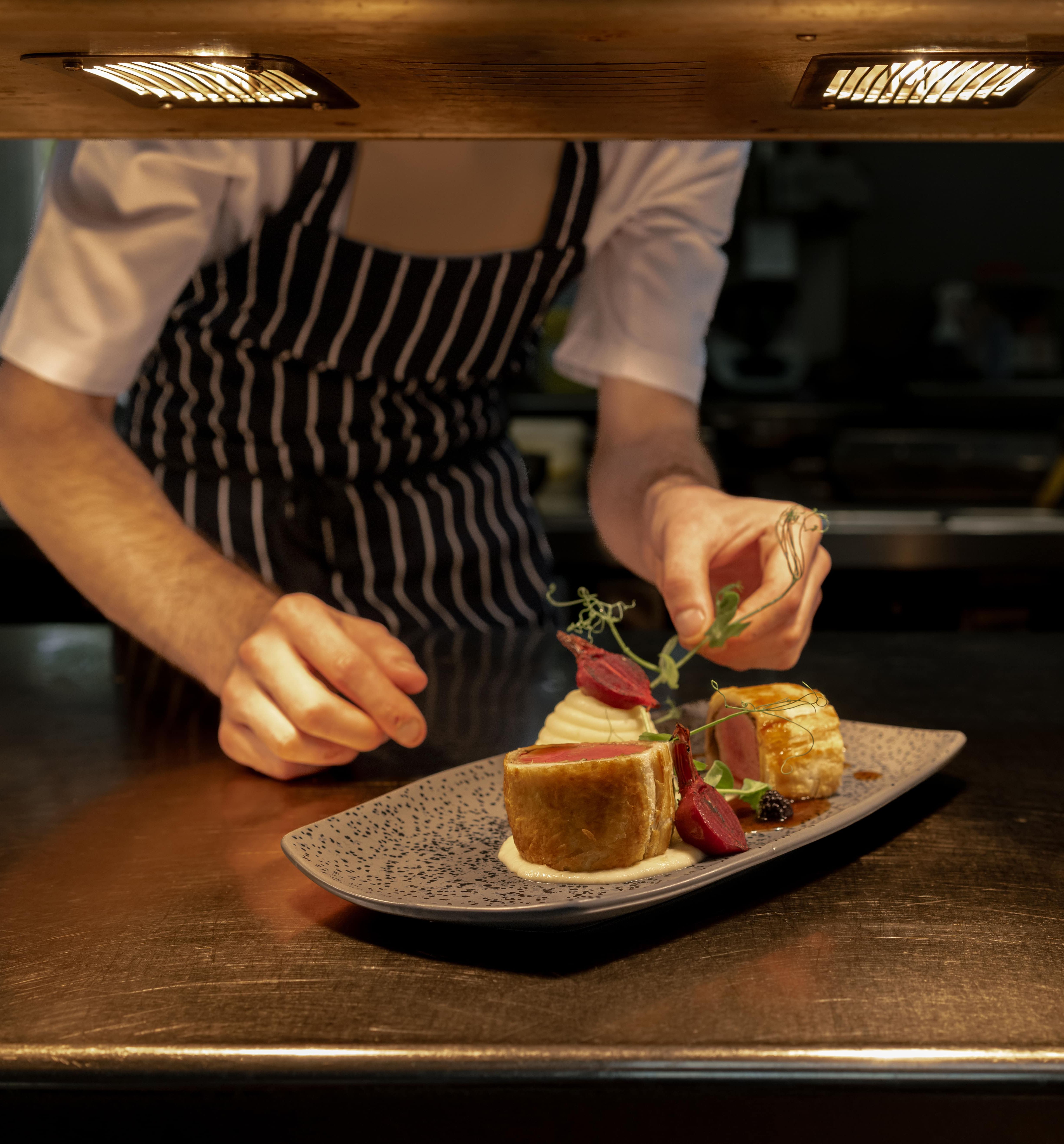 Chef in striped apron carefully garnishing a plated gourmet dish under kitchen lights