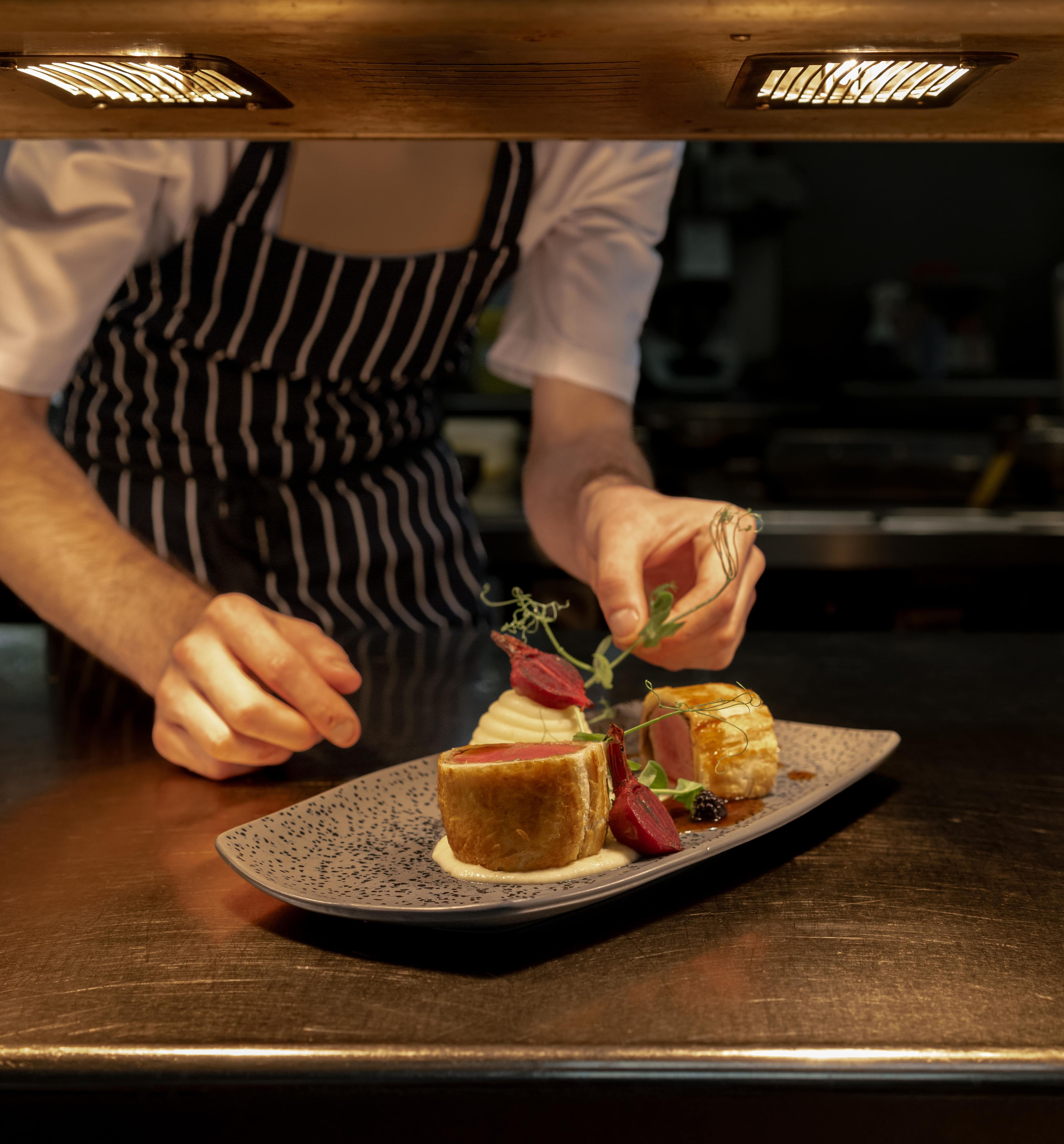 Chef in striped apron carefully garnishing a plated gourmet dish under kitchen lights
