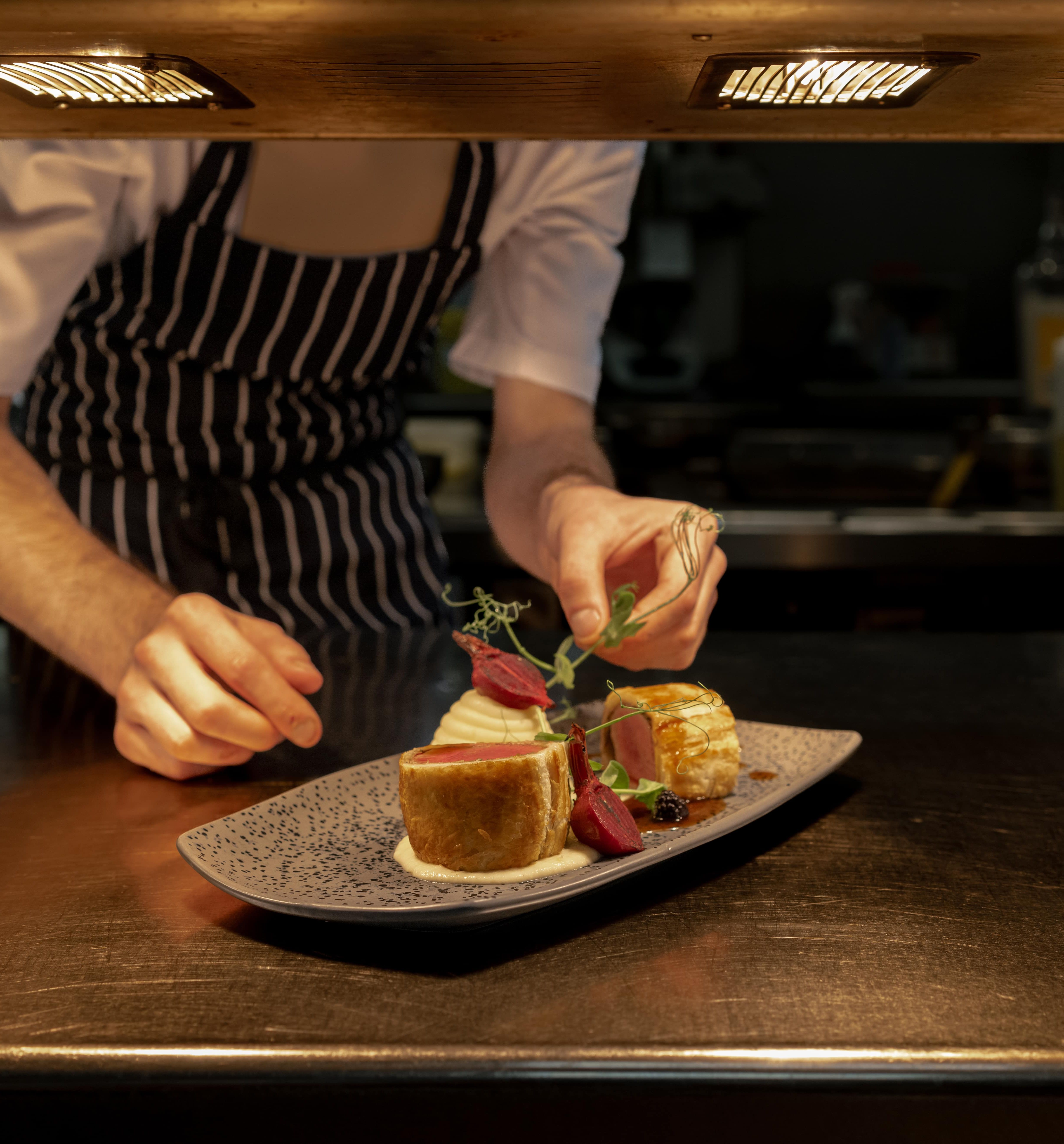 Chef in striped apron carefully garnishing a plated gourmet dish under kitchen lights