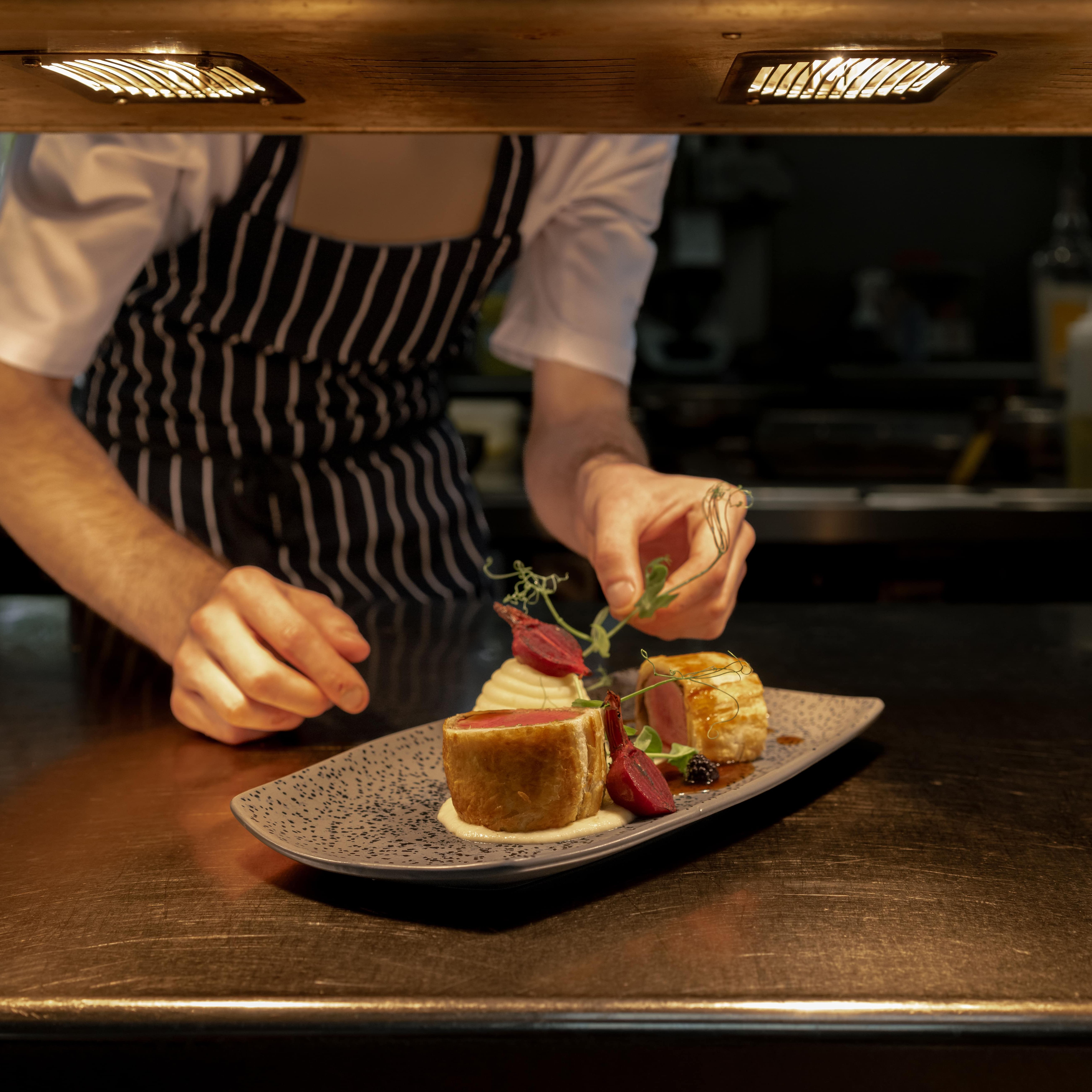 Chef in striped apron carefully garnishing a plated gourmet dish under kitchen lights