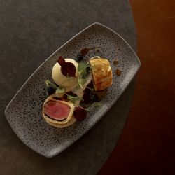 Plated gourmet dish featuring beef Wellington, mashed potatoes, and garnishes on a speckled rectangular plate.