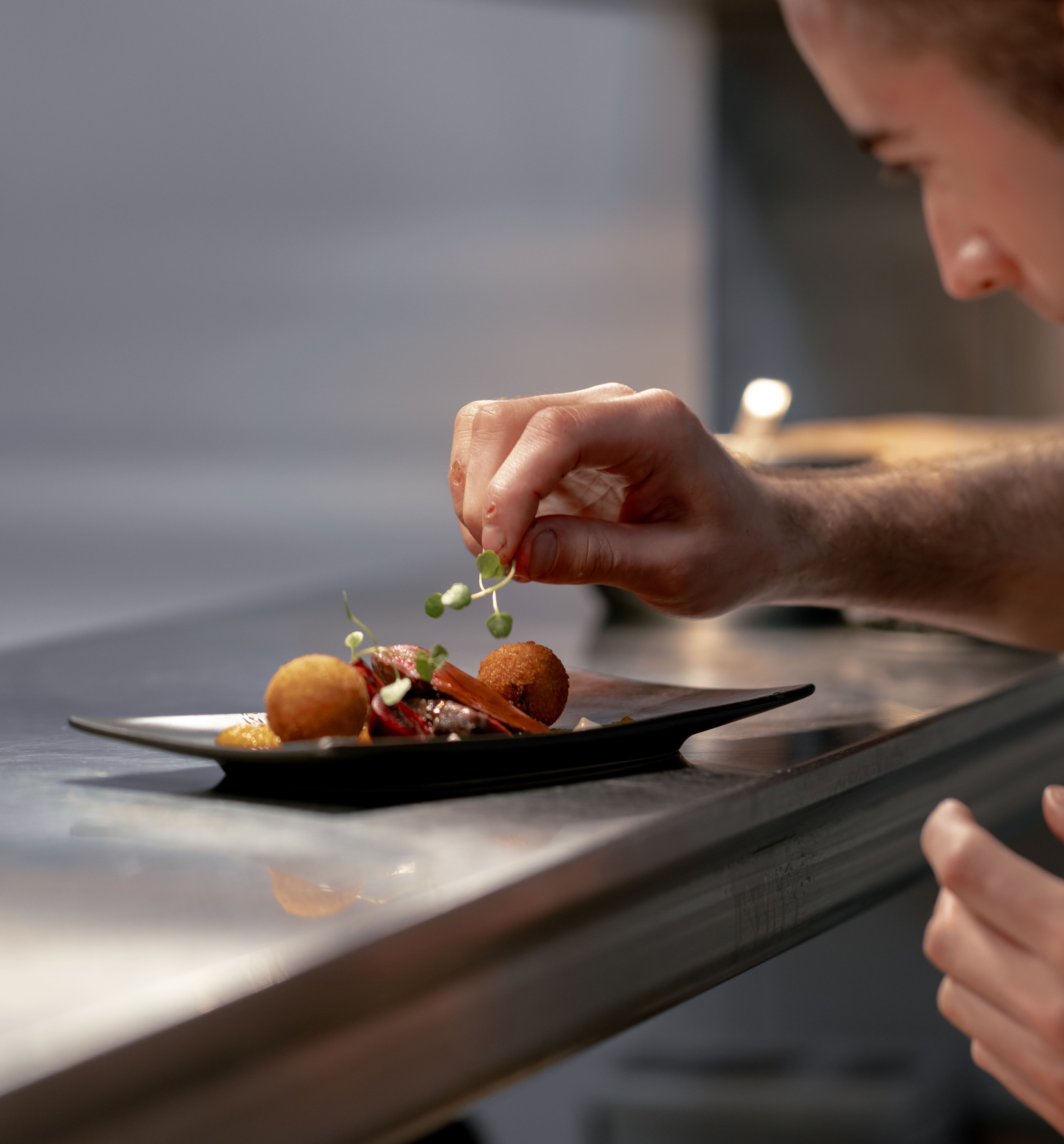 Chef garnishing a plated dish with fresh herbs in a professional kitchen.