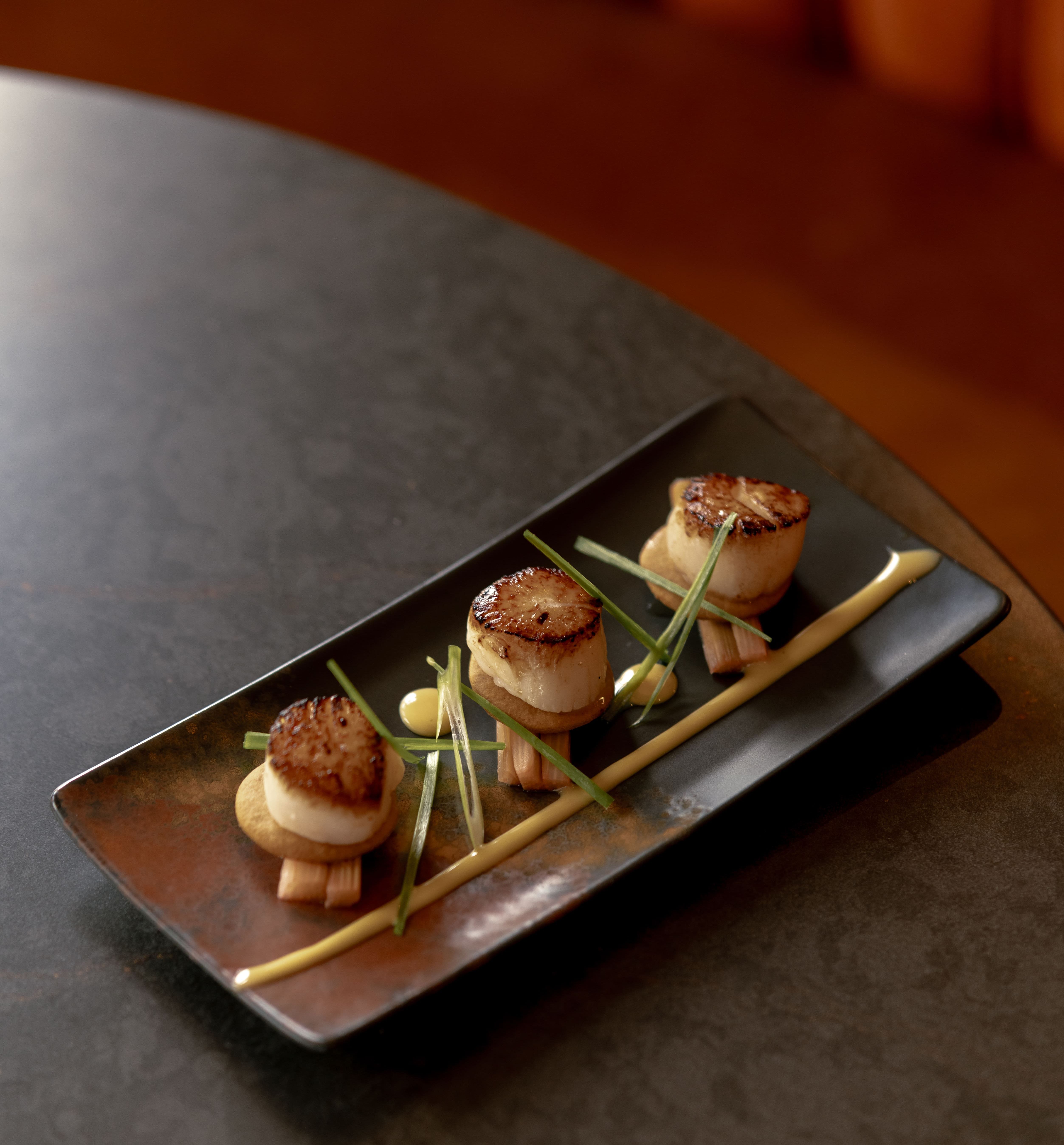 Elegant plate of seared scallops garnished with greens and sauce on a black rectangular dish