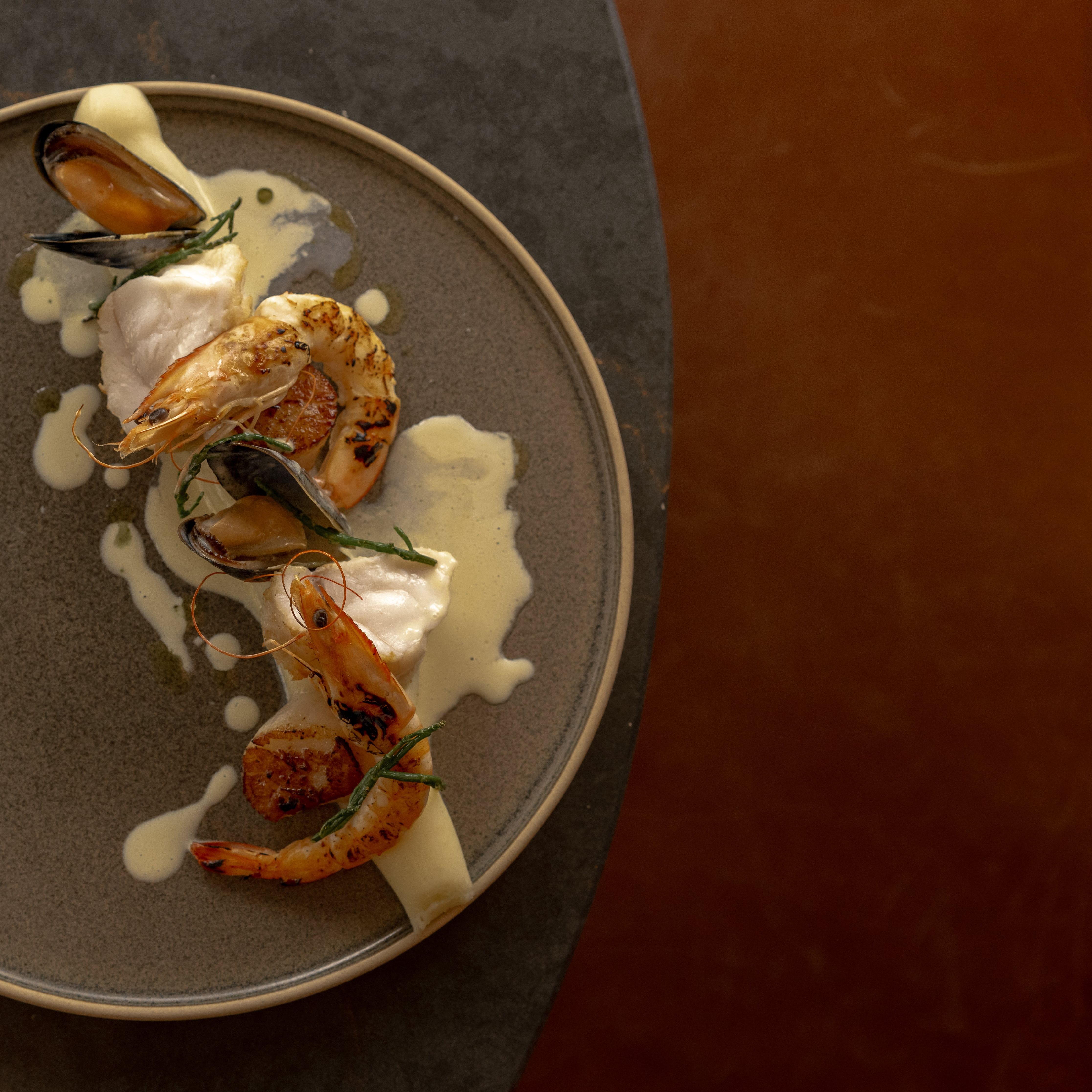 Gourmet seafood dish with shrimp, mussels, and white fish on a gray plate with creamy sauce.