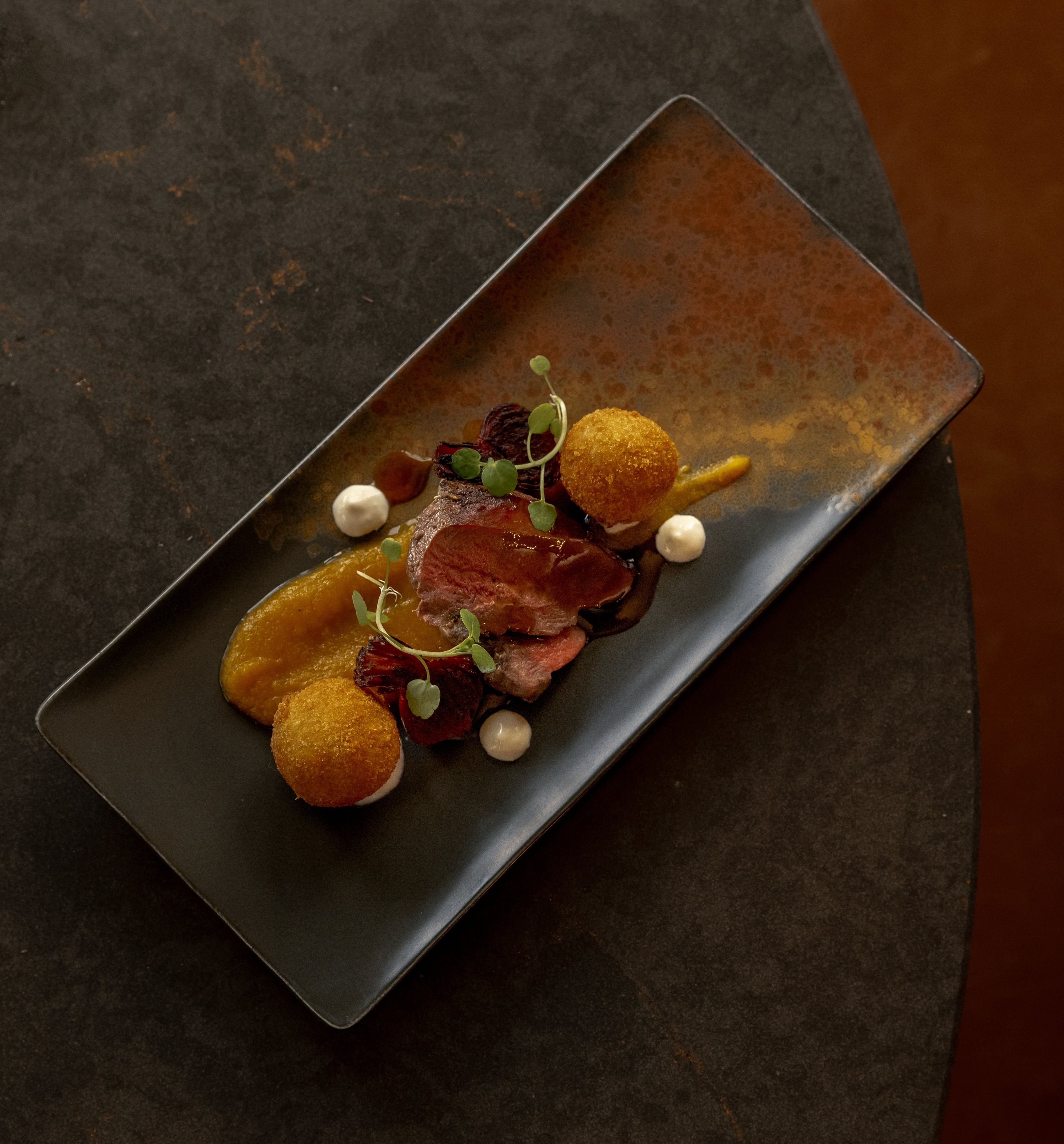 Gourmet plated dish with sliced meat, two golden croquettes, pureed sauce, microgreens, and decorative dollops on a rectangular black plate.