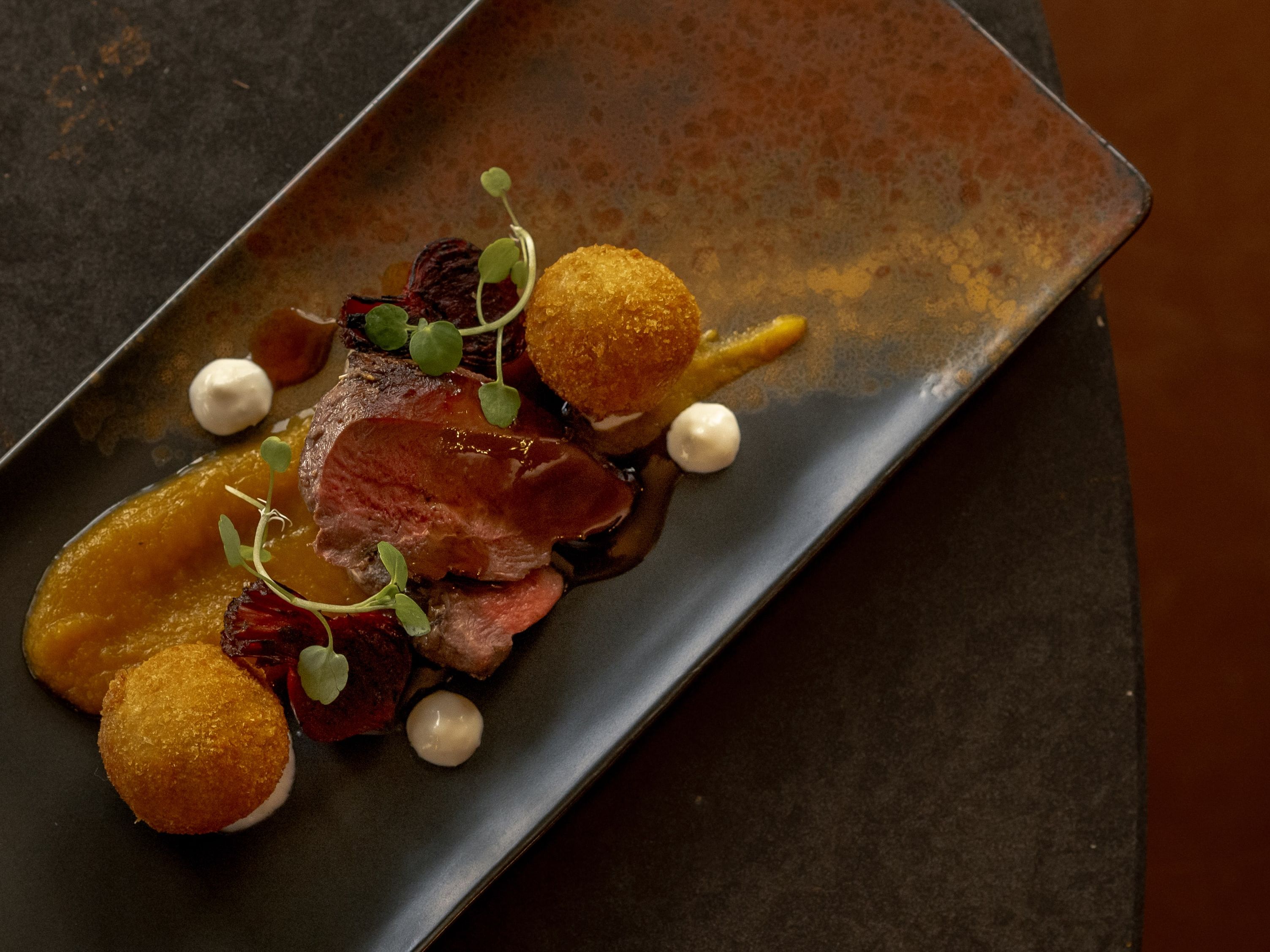 Gourmet plated dish with sliced meat, puree, and croquettes on a dark rectangular plate