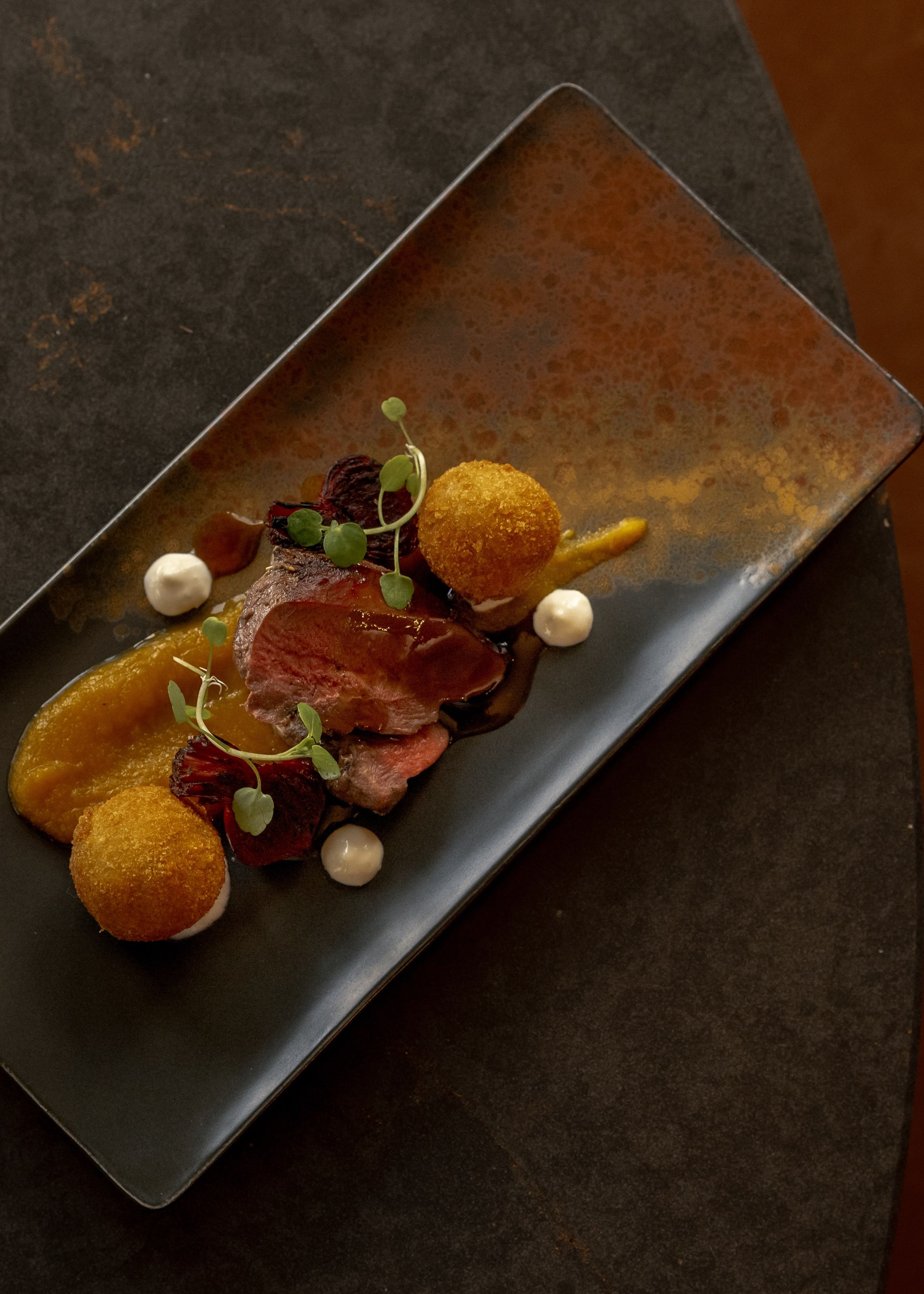 Gourmet plated dish with sliced meat, puree, and croquettes on a dark rectangular plate