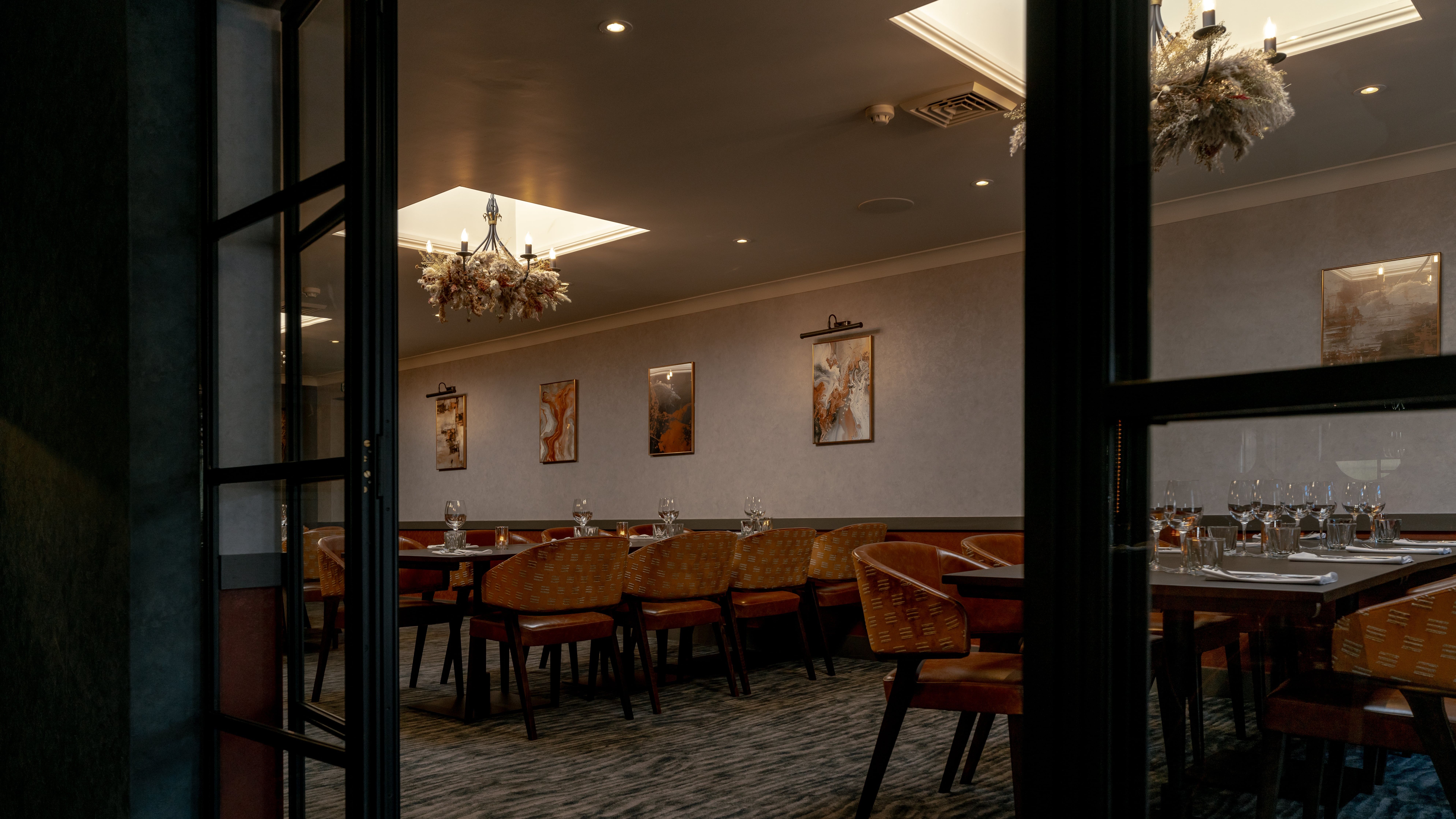 Elegant private dining room with set tables, brown chairs, chandeliers, and artwork on the walls.