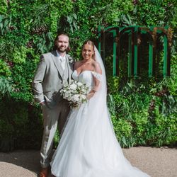 Bride and groom posing together outdoors in front of a lush green wall, bride holding bouquet.