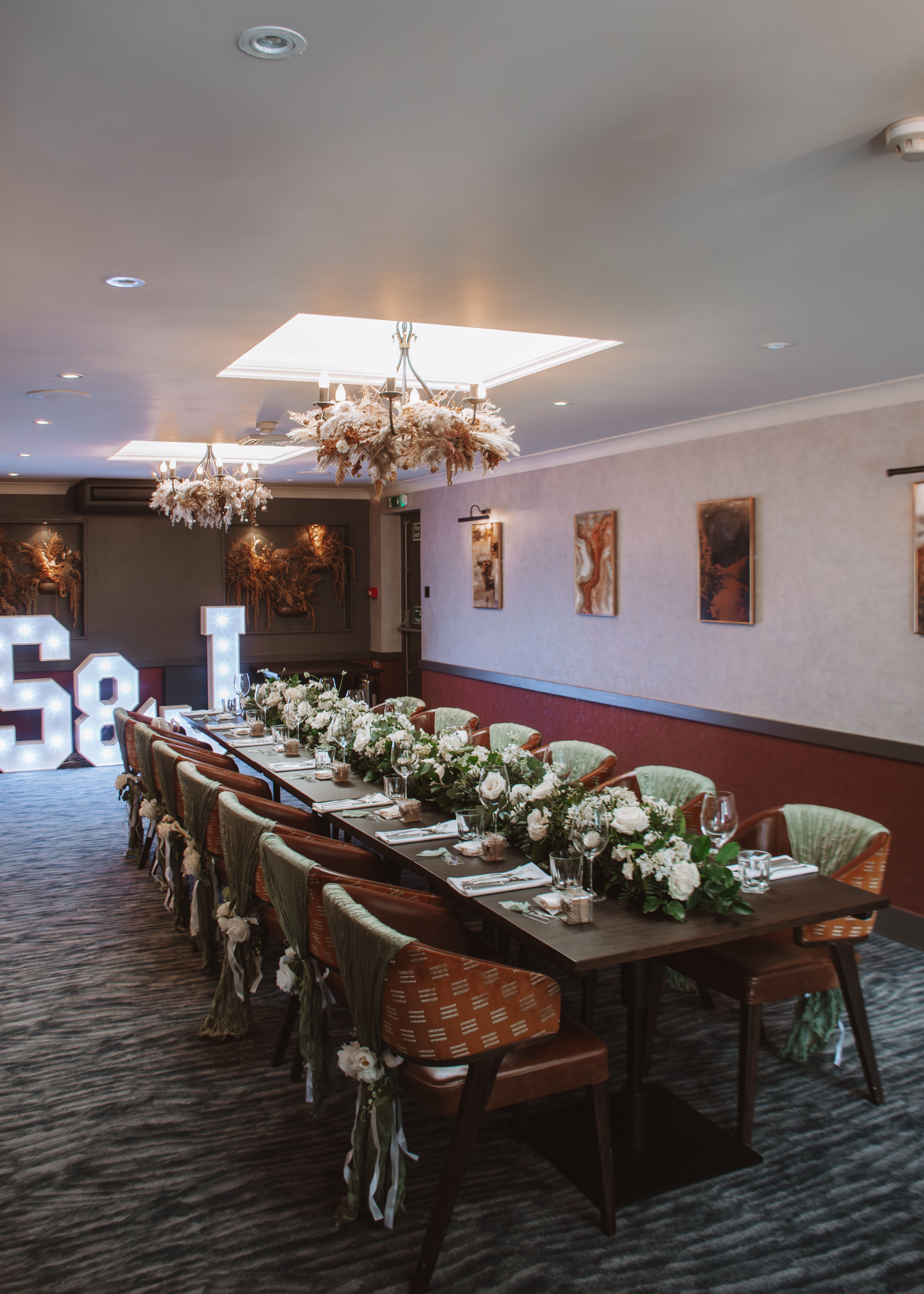 Elegant dining room setup with a long table decorated with white flowers, green fabric on chairs, and illuminated letters in the background.
