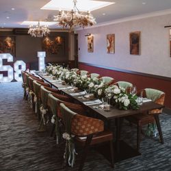 Elegant dining room setup with a long table decorated with white flowers, green fabric on chairs, and illuminated letters in the background.