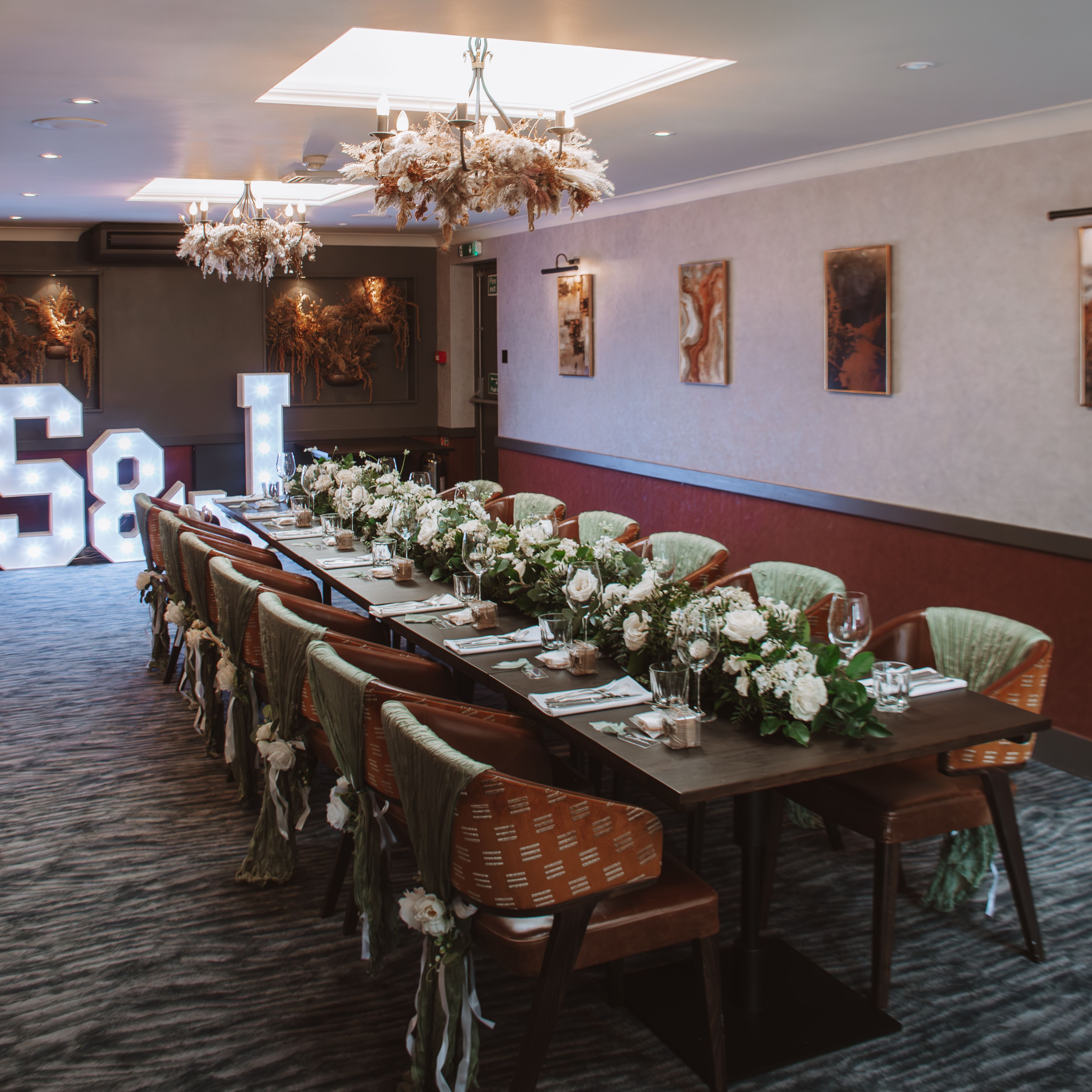 Elegant dining room setup with a long table decorated with white flowers, green fabric on chairs, and illuminated letters in the background.