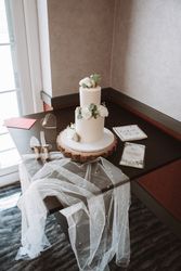 Elegant two-tier white wedding cake decorated with white flowers and greenery, sitting on a wooden cake stand.