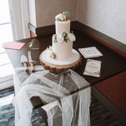 Elegant two-tier white wedding cake decorated with white flowers and greenery, sitting on a wooden cake stand.