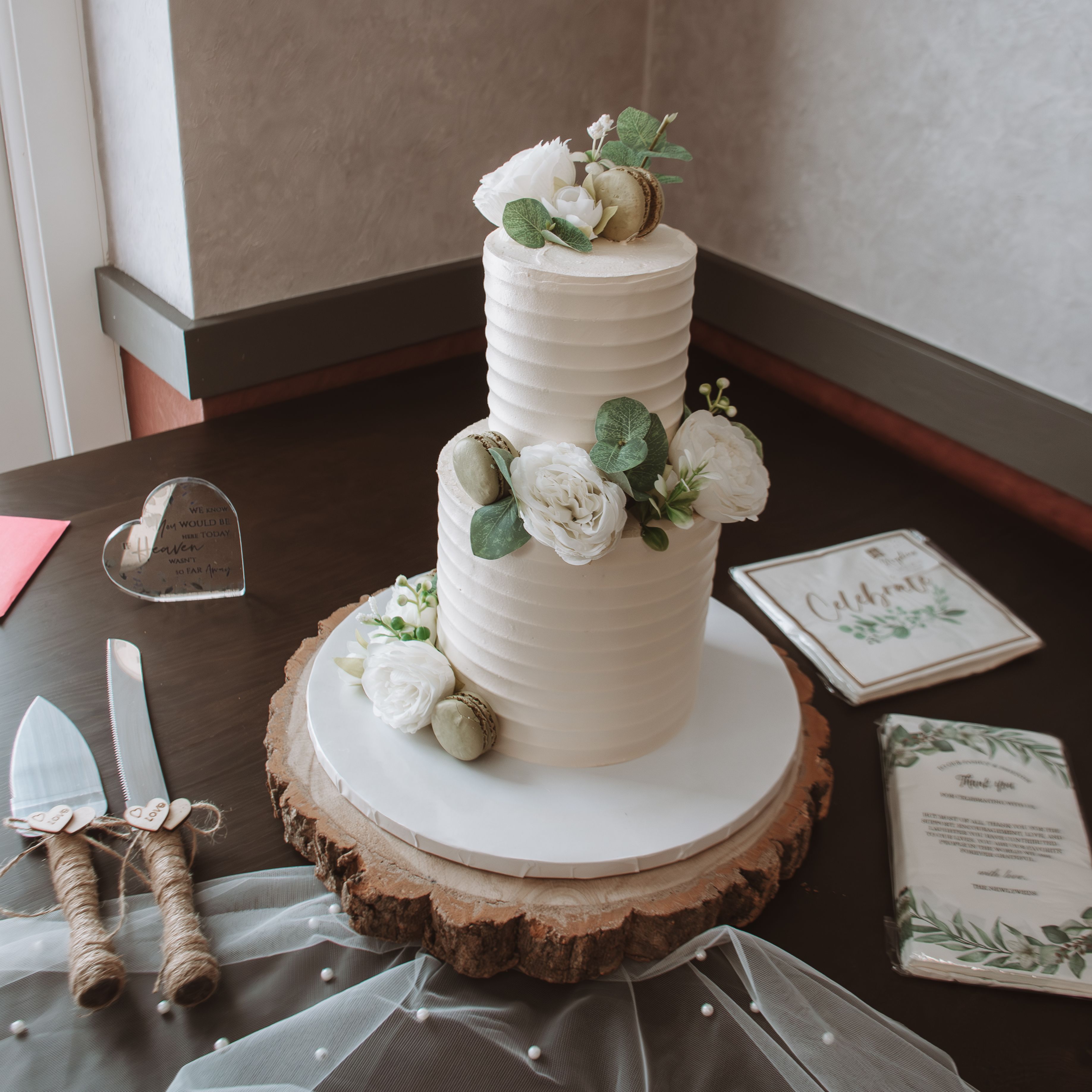 Two-tiered white wedding cake decorated with white flowers and macarons, placed on a wooden cake stand.