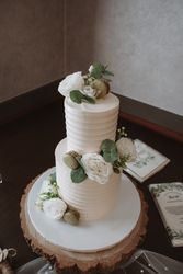 Two-tiered white wedding cake decorated with white flowers, green leaves, and macarons, placed on a wooden cake stand.