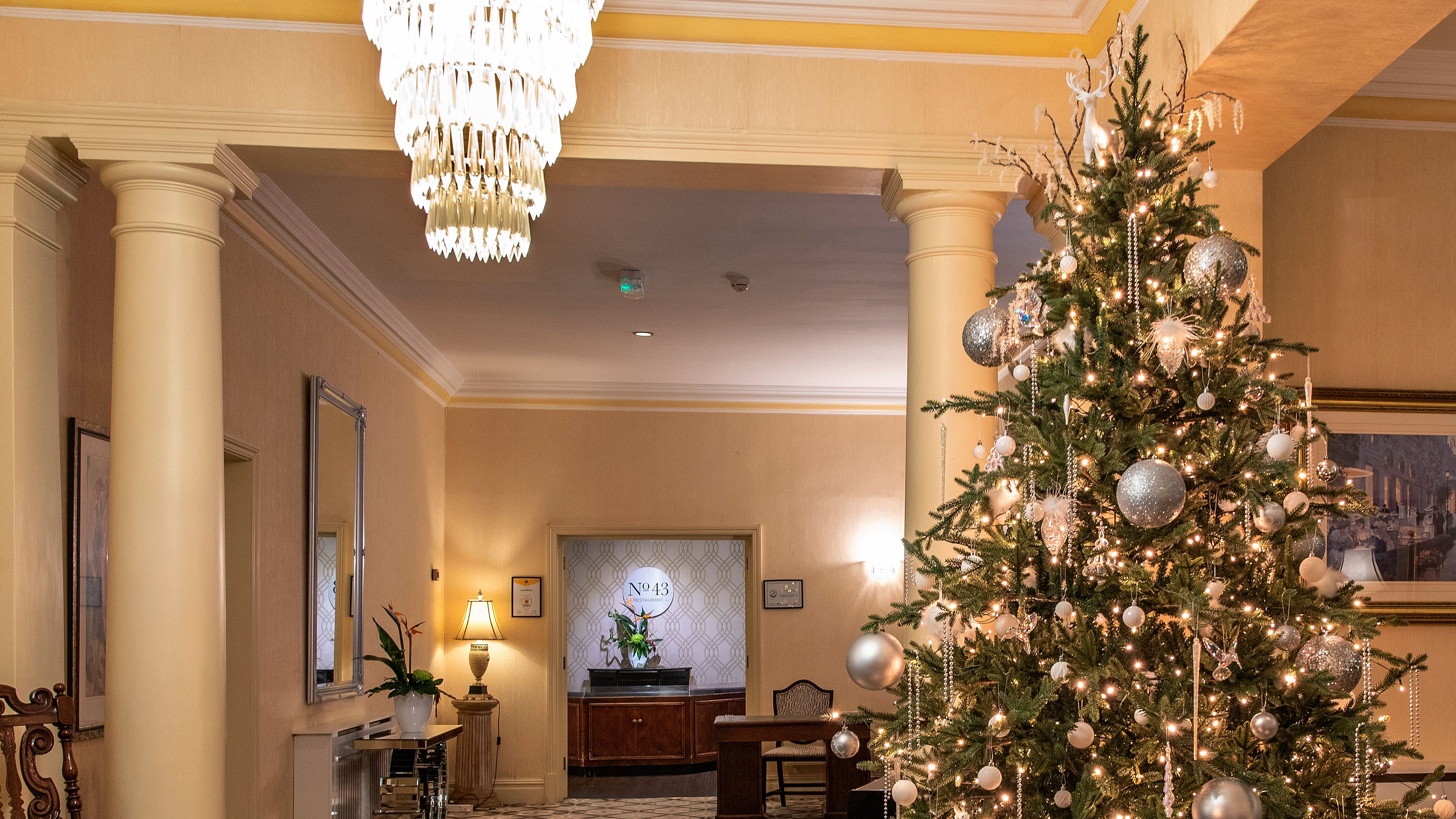 Elegant hotel lobby decorated for Christmas with a tall, festive tree adorned with silver ornaments and lights, surrounded by wrapped gifts, under a sparkling chandelier.