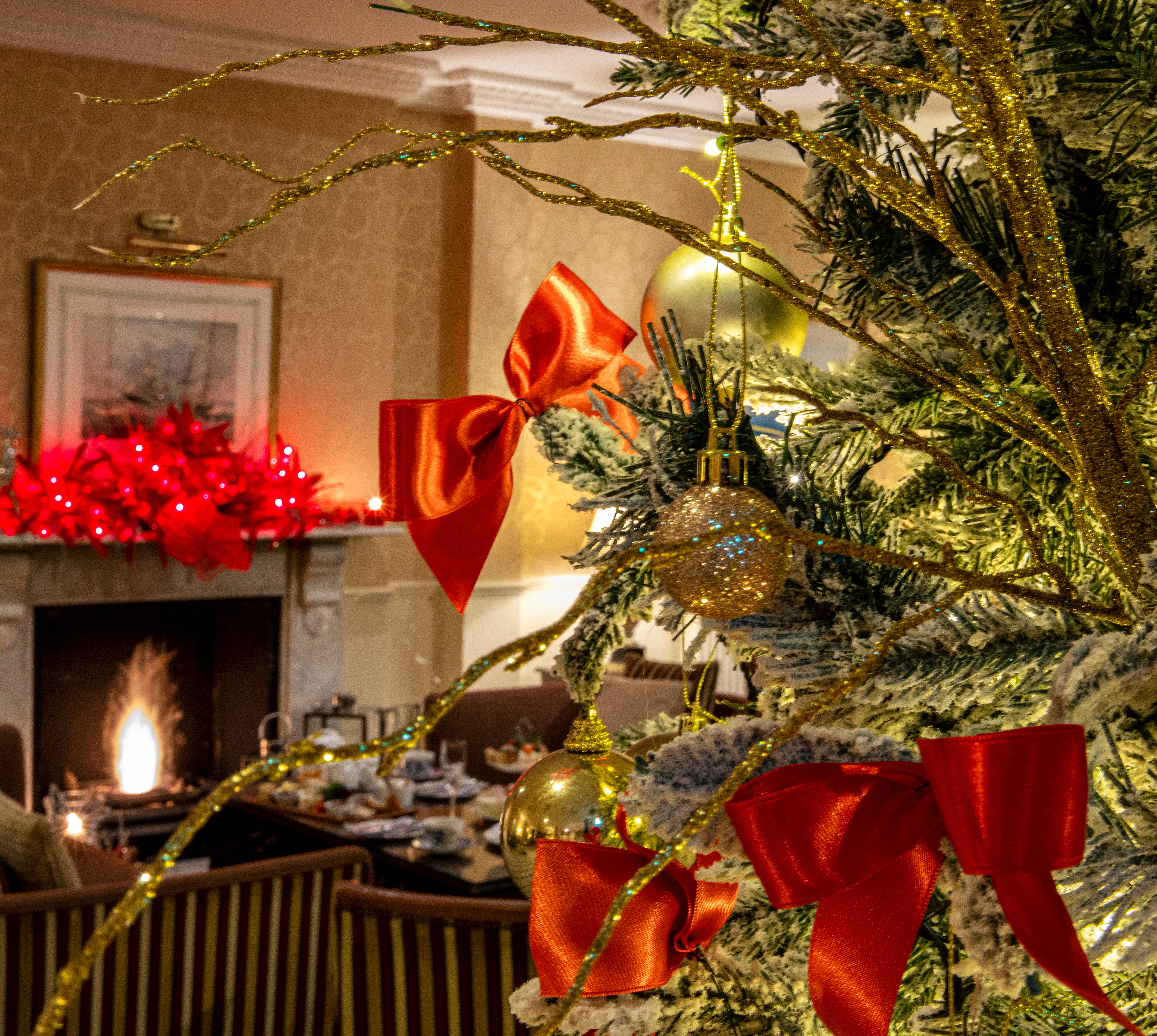 Close-up of a decorated Christmas tree with red bows and gold ornaments, set in a cozy lounge with a festive fireplace and a warmly lit holiday display in the background.