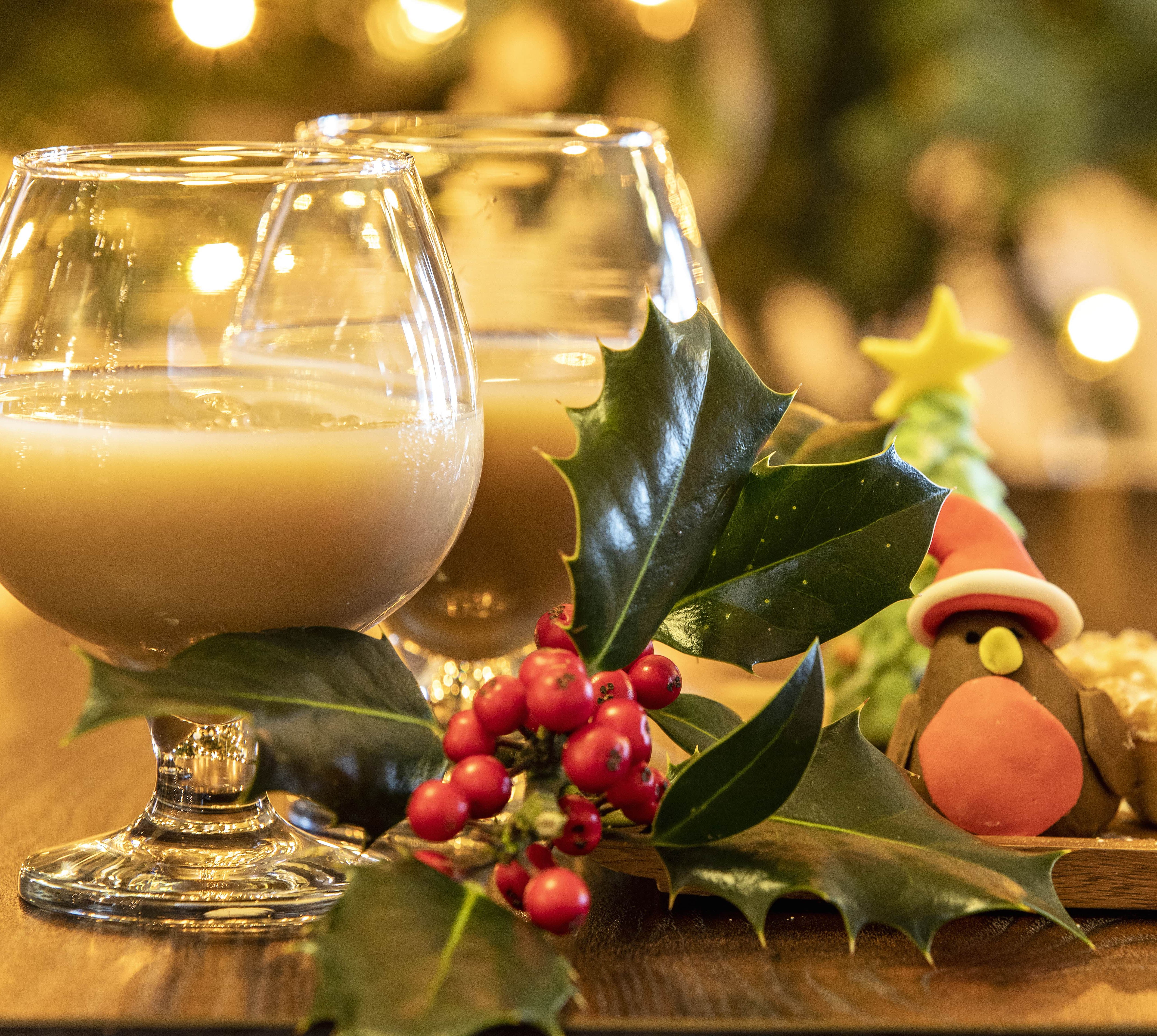 Festive holiday setup with two glasses of eggnog, holly sprigs, a decorated Christmas cookie shaped like a robin, and mince pies, set against a warm, twinkling backdrop.