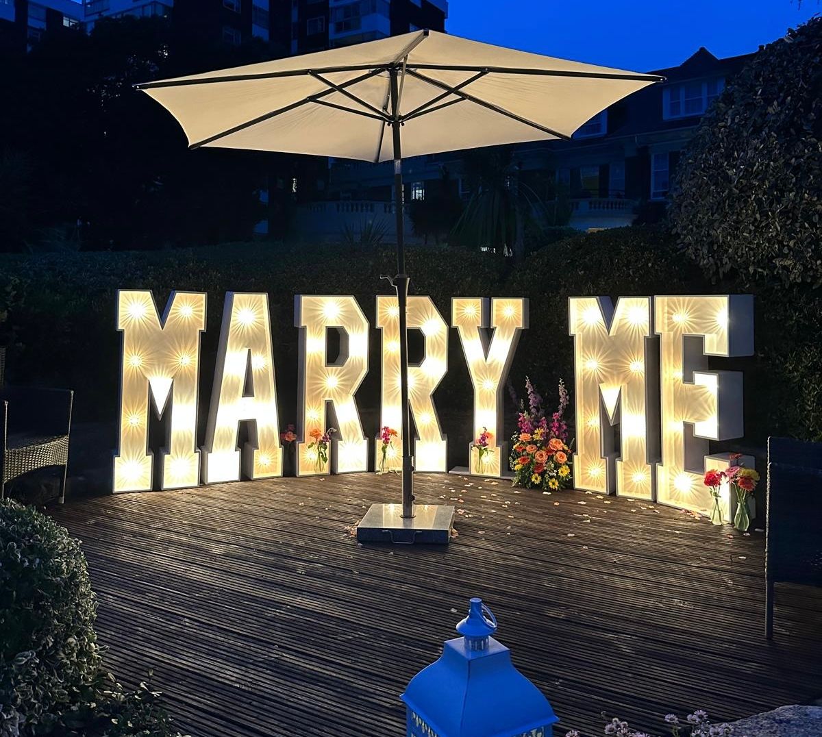Large illuminated letters spelling 'Marry Me' under an outdoor umbrella at night, with flowers and a decorative lantern nearby.