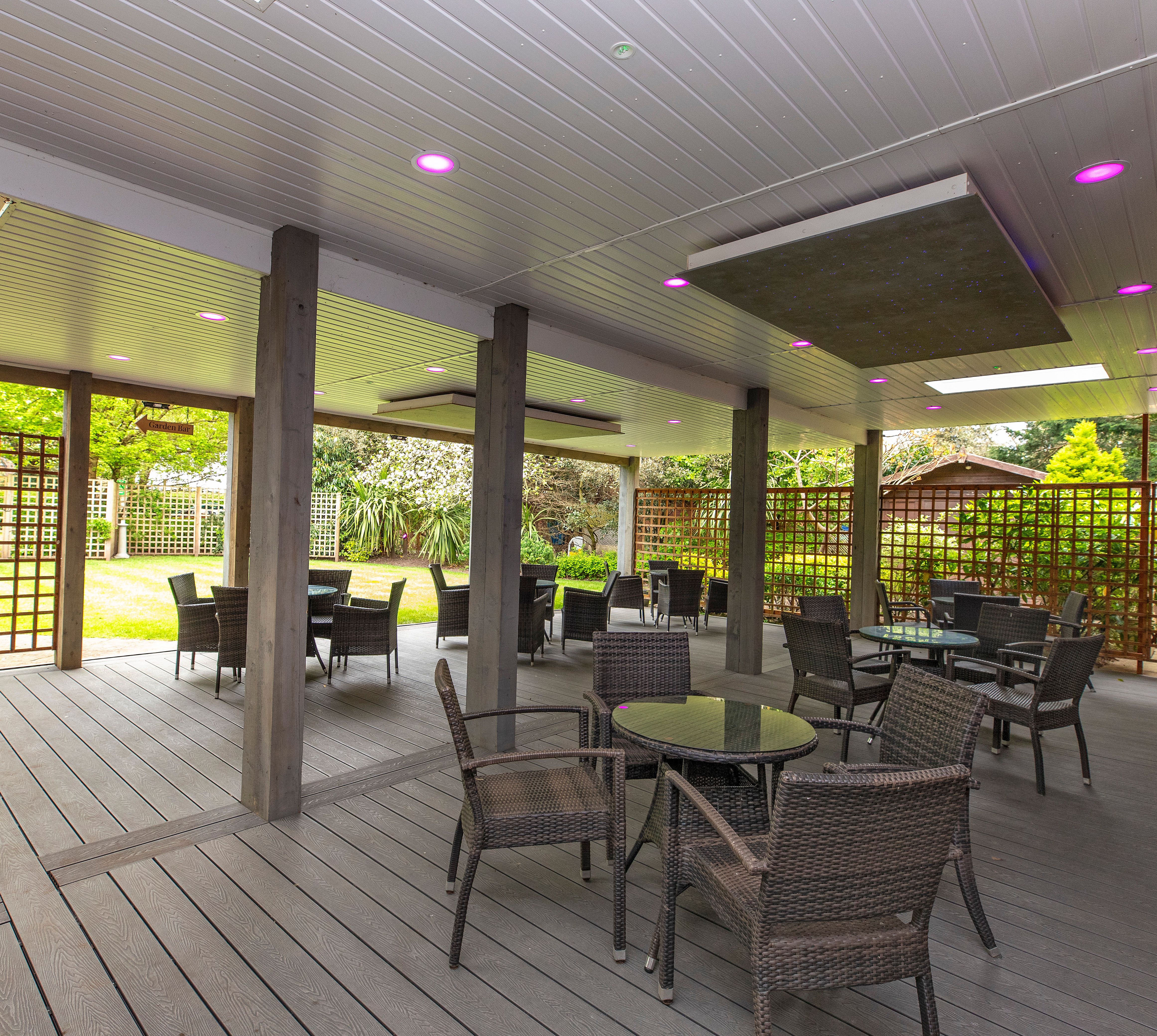 Covered outdoor patio with wicker chairs and glass-top tables, grey decking and purple recessed ceiling lights