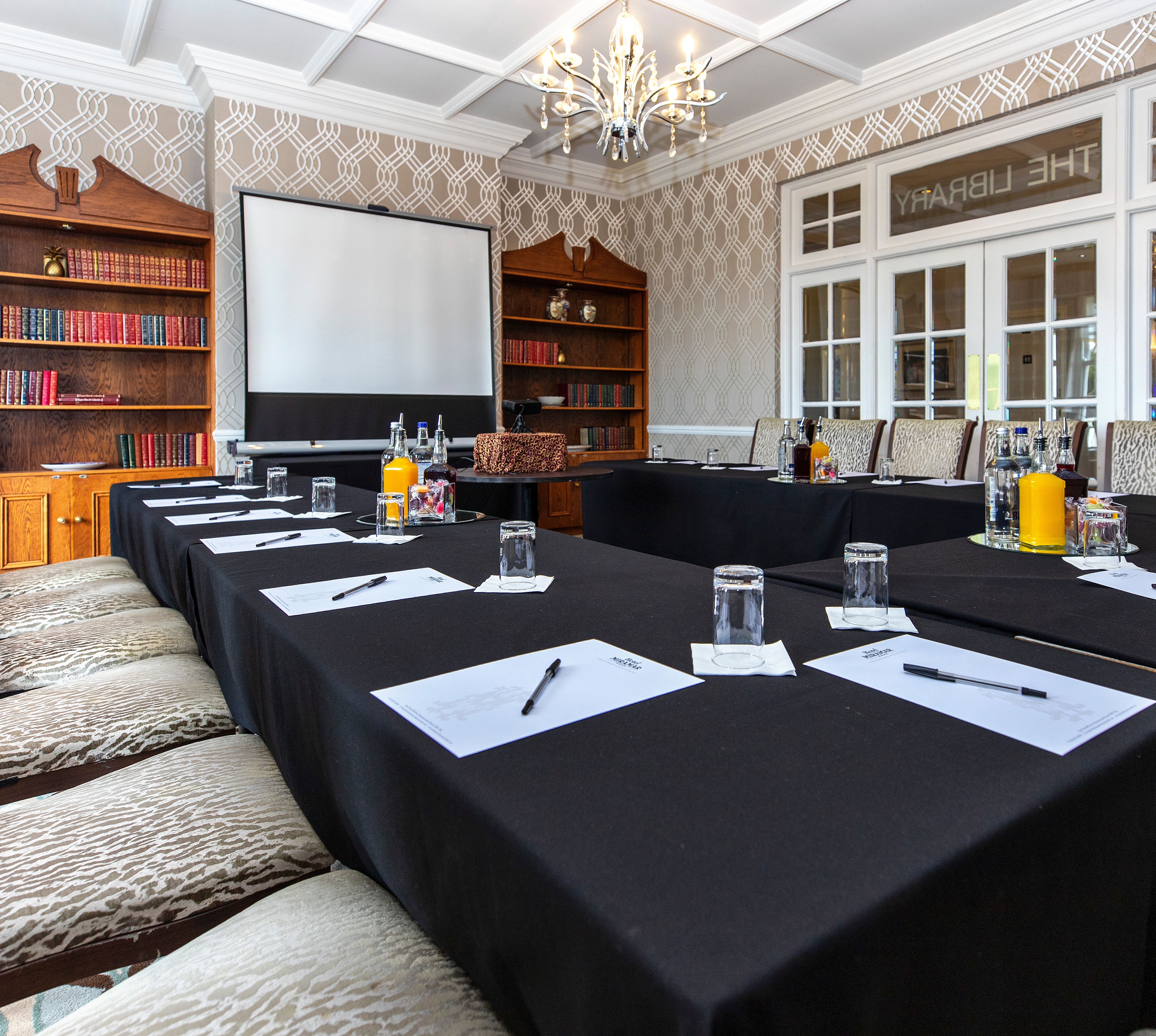 Boardroom-style meeting room with U-shaped tables, notepads, water glasses and a projector screen beside bookcases