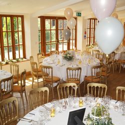 A bright and elegant wedding reception setup in a sunlit room with round tables covered in white tablecloths, gold chairs, and neatly arranged glassware.