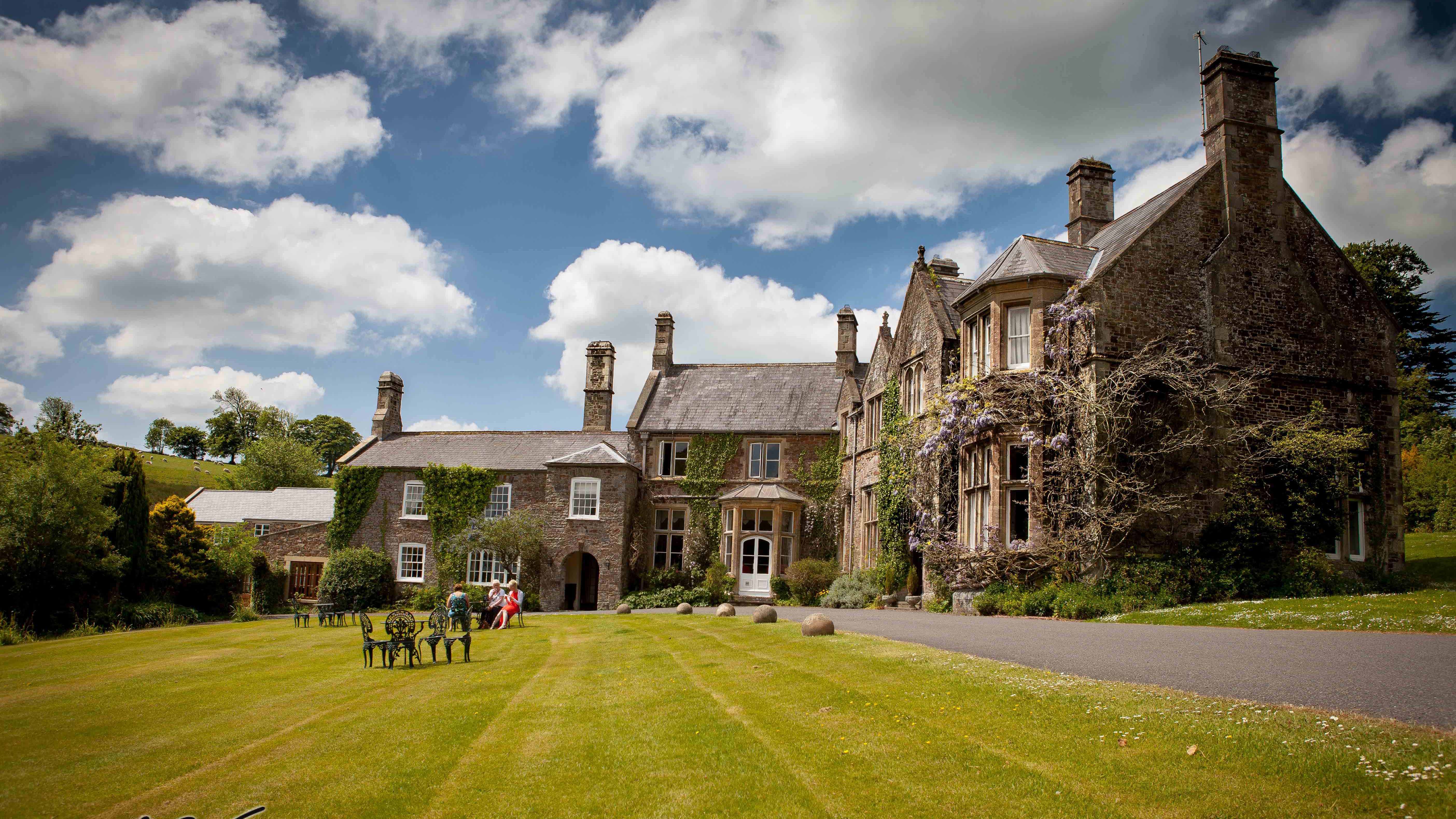 stately manor house with ivy-covered stone walls and tall chimneys sits under a partly cloudy sky.