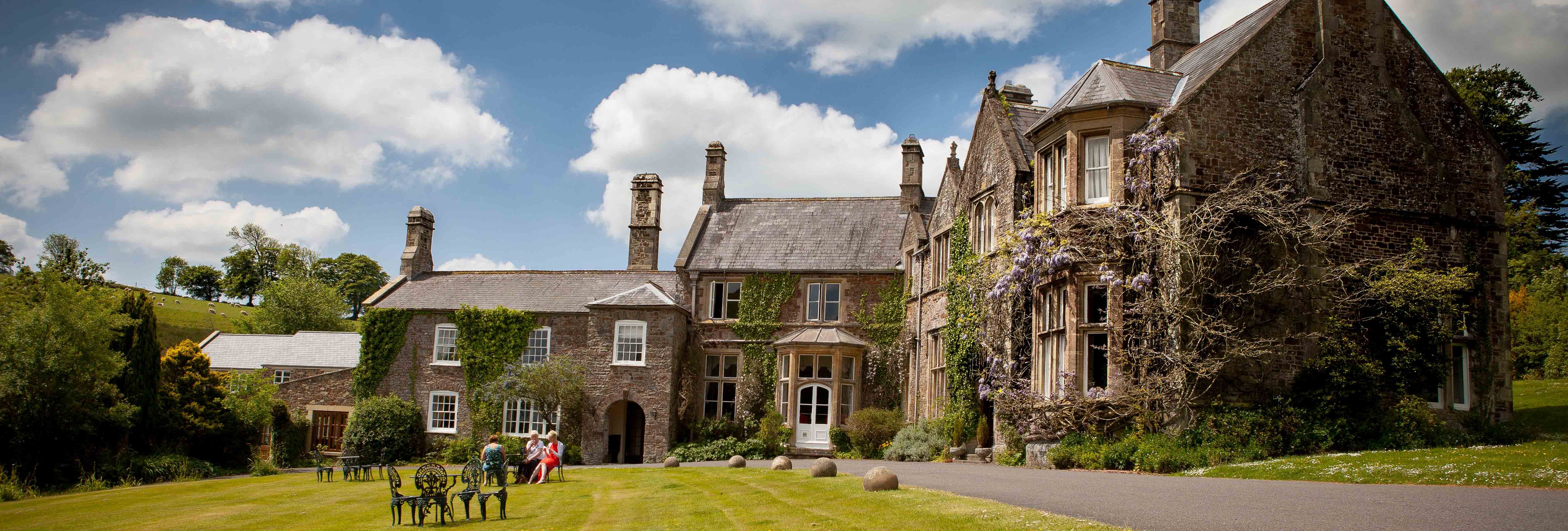 stately manor house with ivy-covered stone walls and tall chimneys sits under a partly cloudy sky.