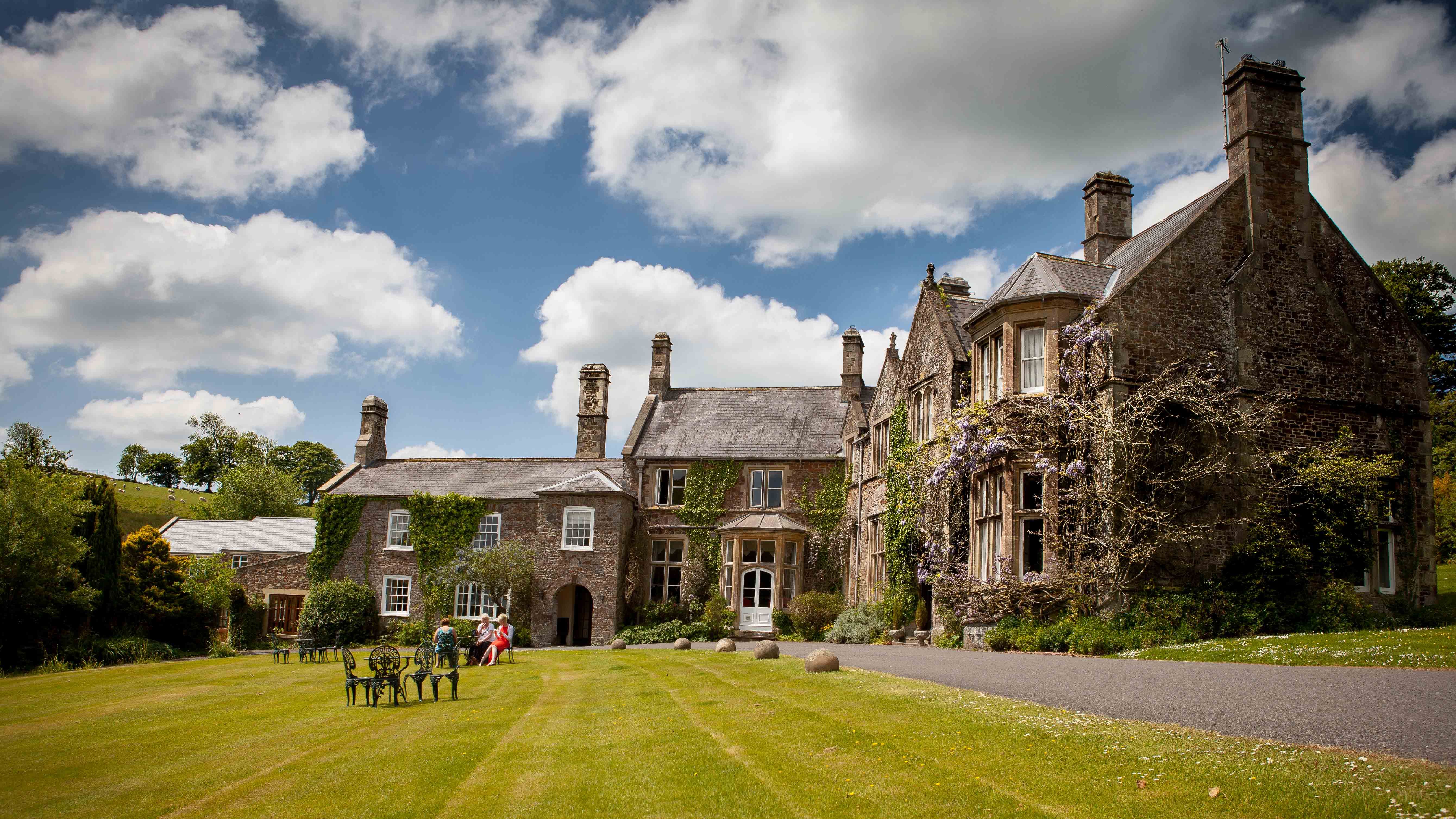 stately manor house with ivy-covered stone walls and tall chimneys sits under a partly cloudy sky.