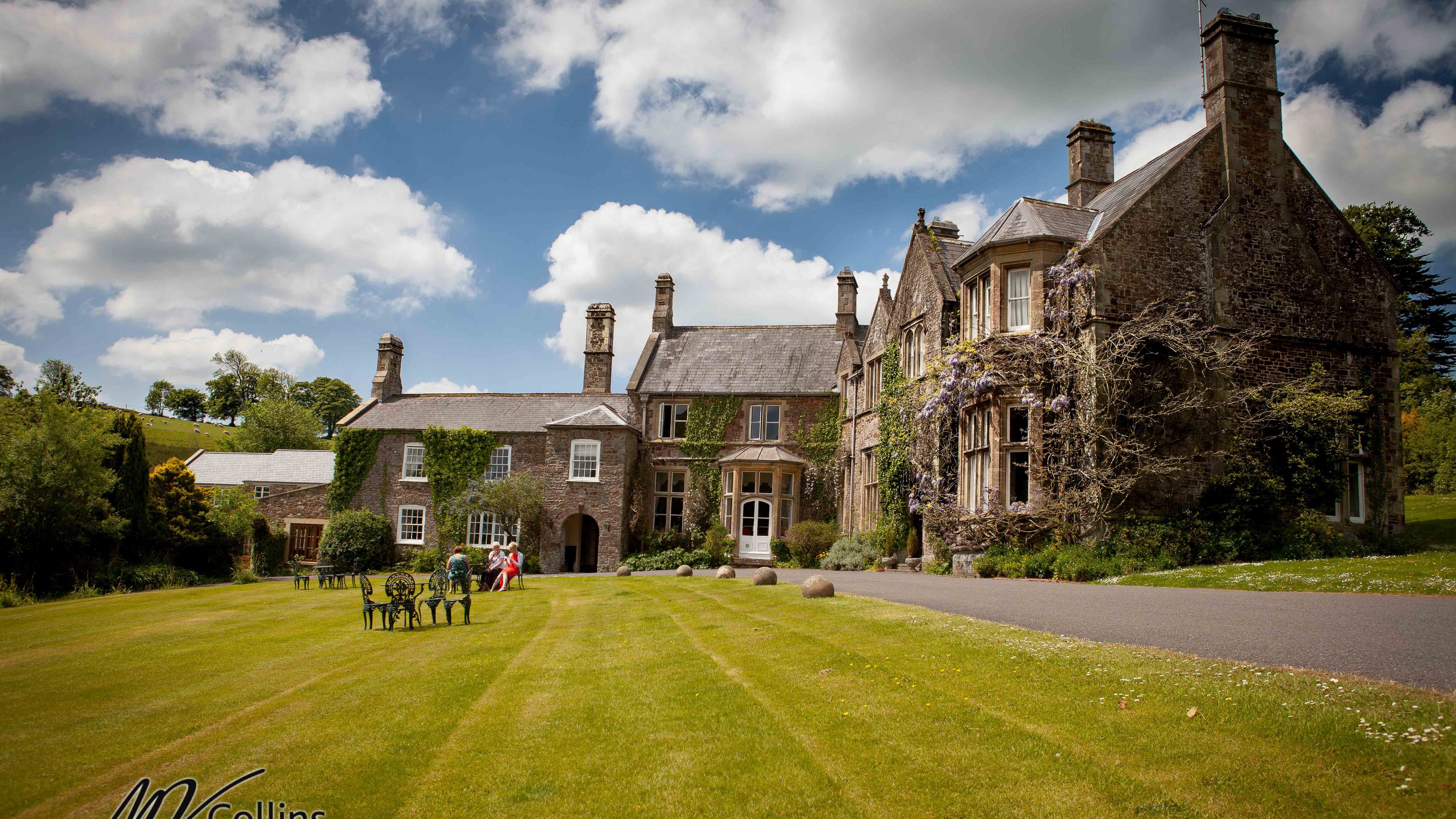 stately manor house with ivy-covered stone walls and tall chimneys sits under a partly cloudy sky.
