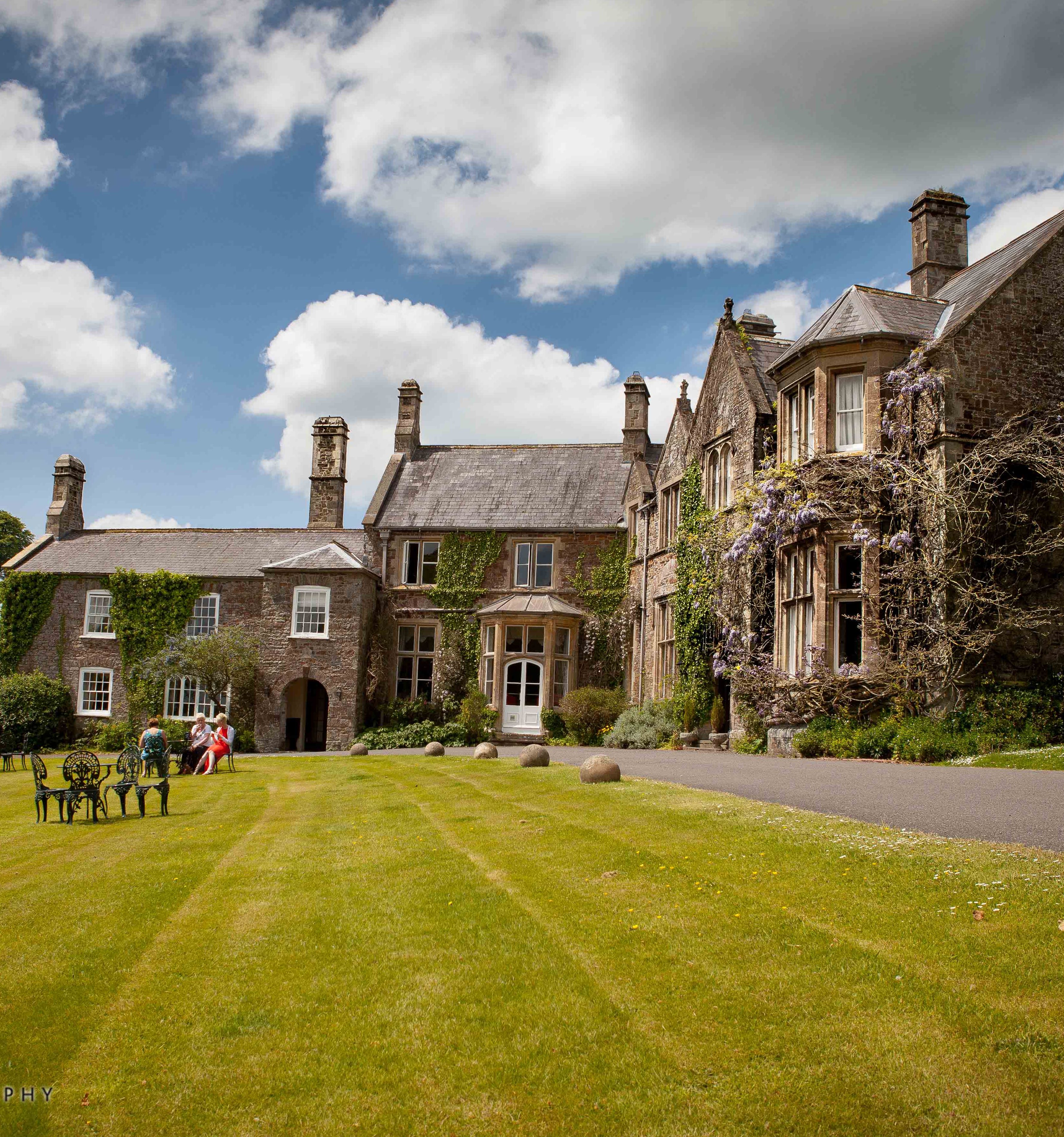 stately manor house with ivy-covered stone walls and tall chimneys sits under a partly cloudy sky.
