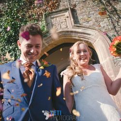 A smiling bride and groom walk hand-in-hand out of a stone archway as guests shower them with colorful flower petals