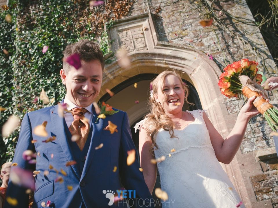 A smiling bride and groom walk hand-in-hand out of a stone archway as guests shower them with colorful flower petals