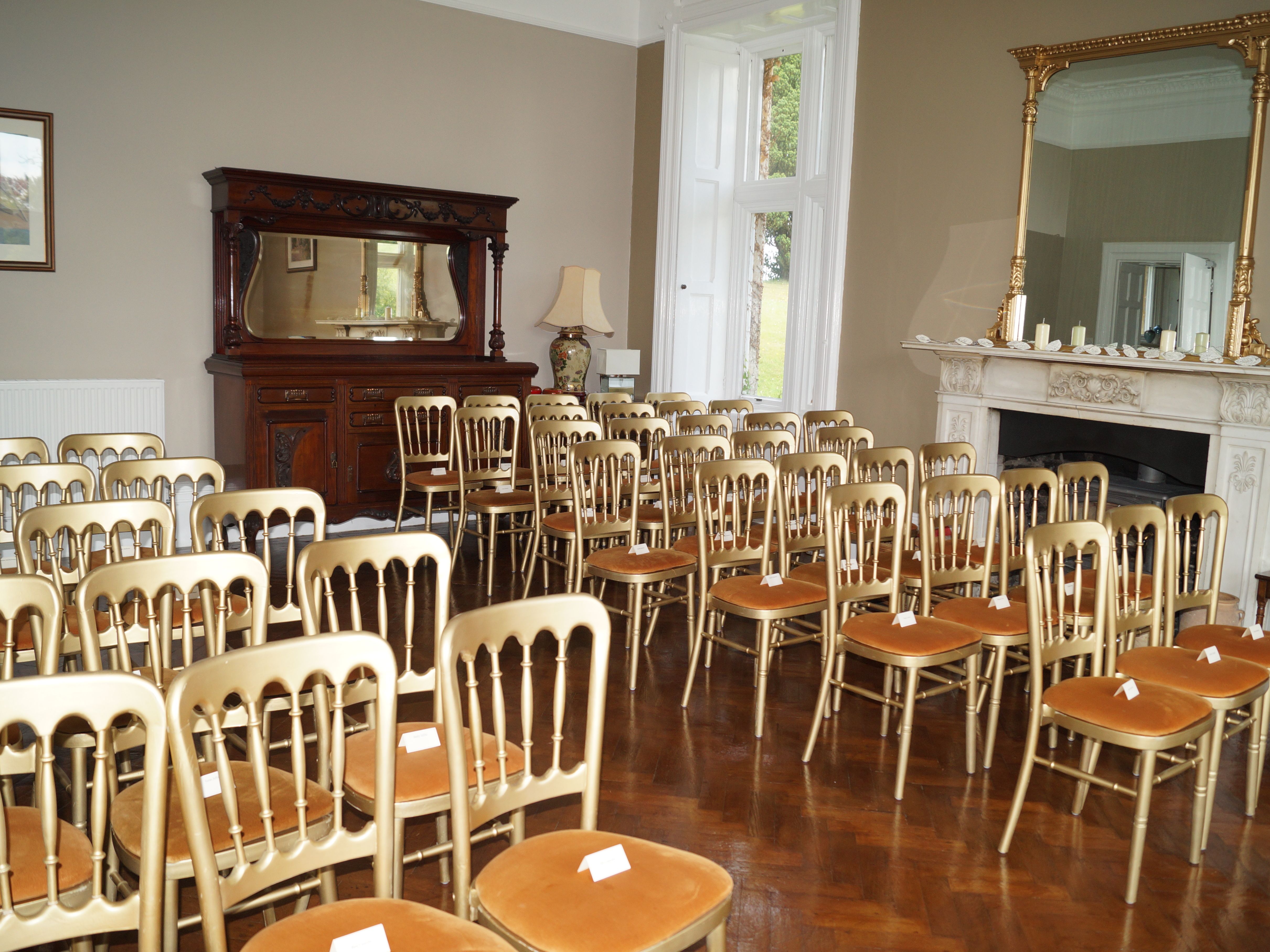 legant room set up for a wedding ceremony, featuring rows of gold chairs with cushioned seats neatly arranged facing a fireplace with a large ornate mirror.