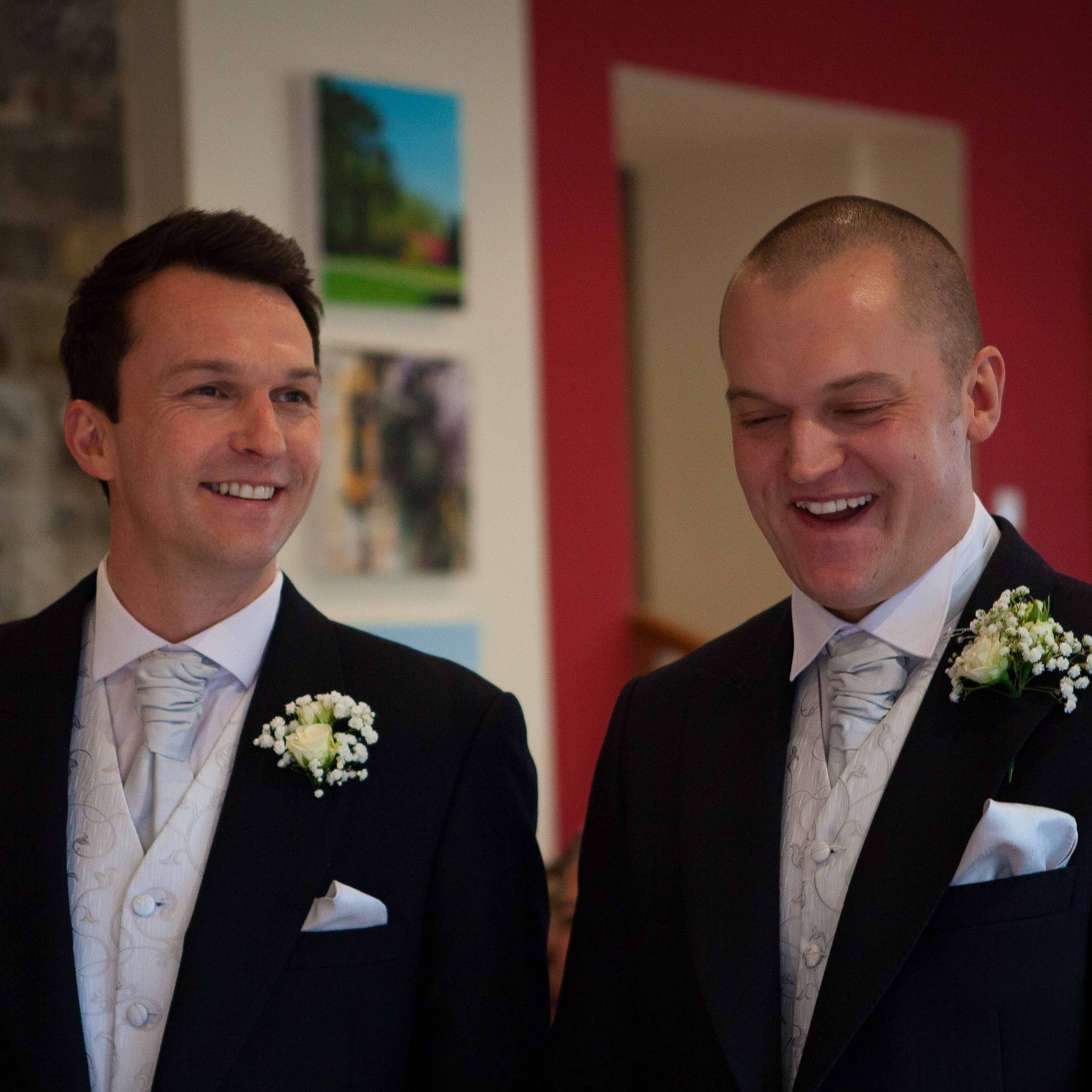Two groomsmen in matching black suits, white vests, and light silver ties stand together, smiling and laughing