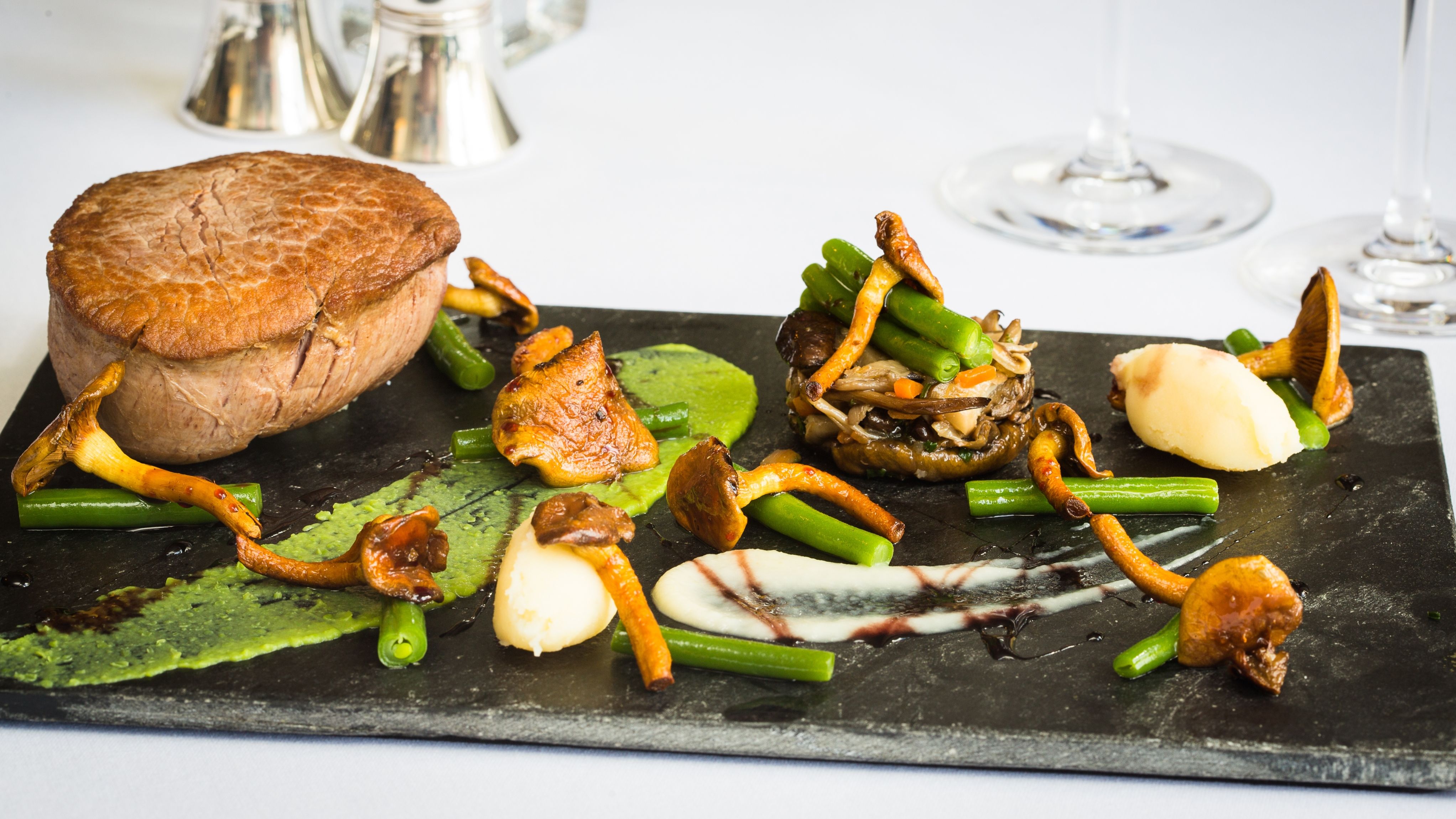 A gourmet dish presented on a dark slate plate, featuring a seared fillet of beef, sautéed wild mushrooms,