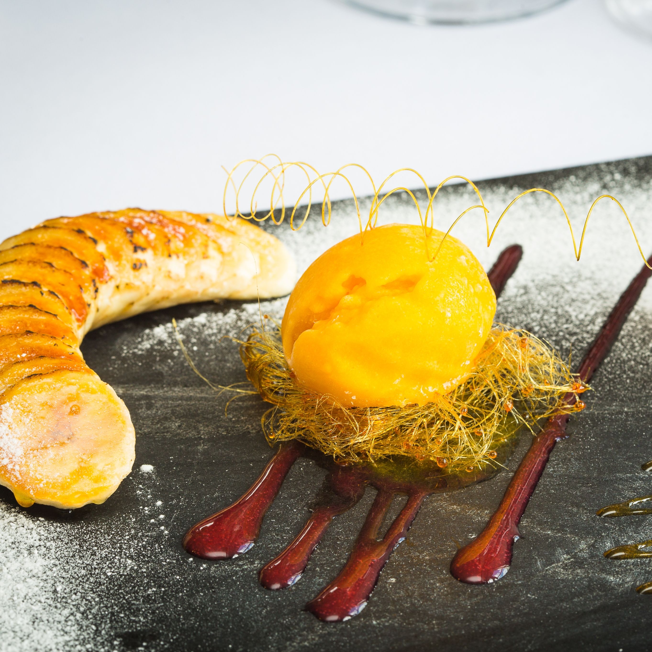 A gourmet dessert plate featuring a caramelized sliced banana, a scoop of mango sorbet nestled in a delicate caramel nest