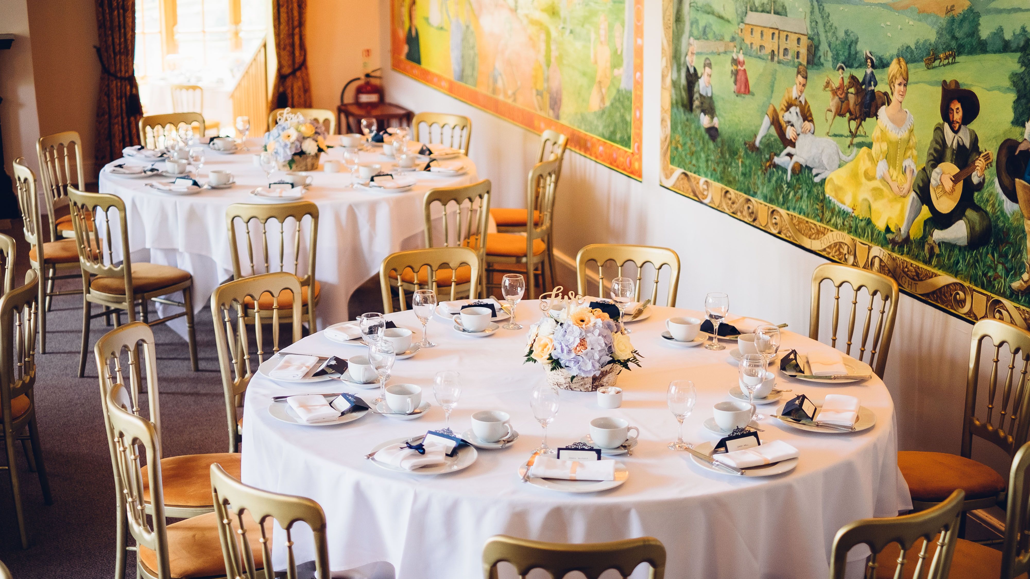 A dining area set for an event with round tables covered in white tablecloths, gold chairs, and elegant place settings.