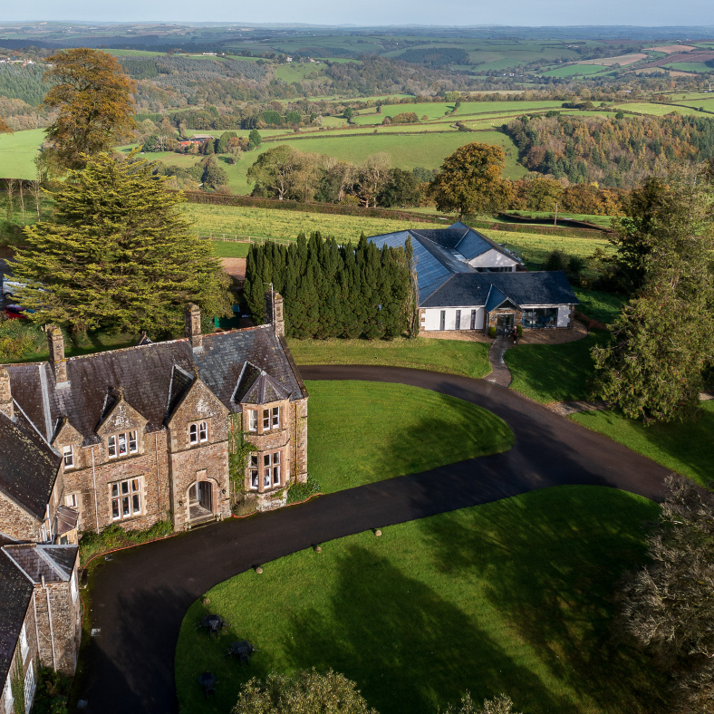 Aerial view of a large countryside estate with historic stone buildings, surrounded by gardens, trees, and rolling green fields.