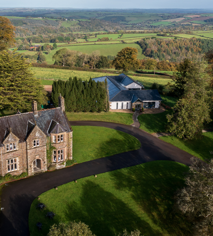 Aerial view of a large countryside estate with historic stone buildings, surrounded by gardens, trees, and rolling green fields.