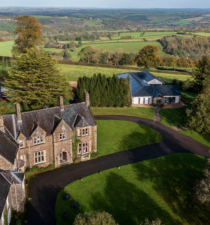 Aerial view of a large countryside estate with historic stone buildings, surrounded by gardens, trees, and rolling green fields.