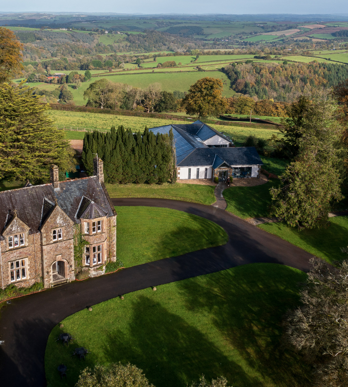 Aerial view of a large countryside estate with historic stone buildings, surrounded by gardens, trees, and rolling green fields.