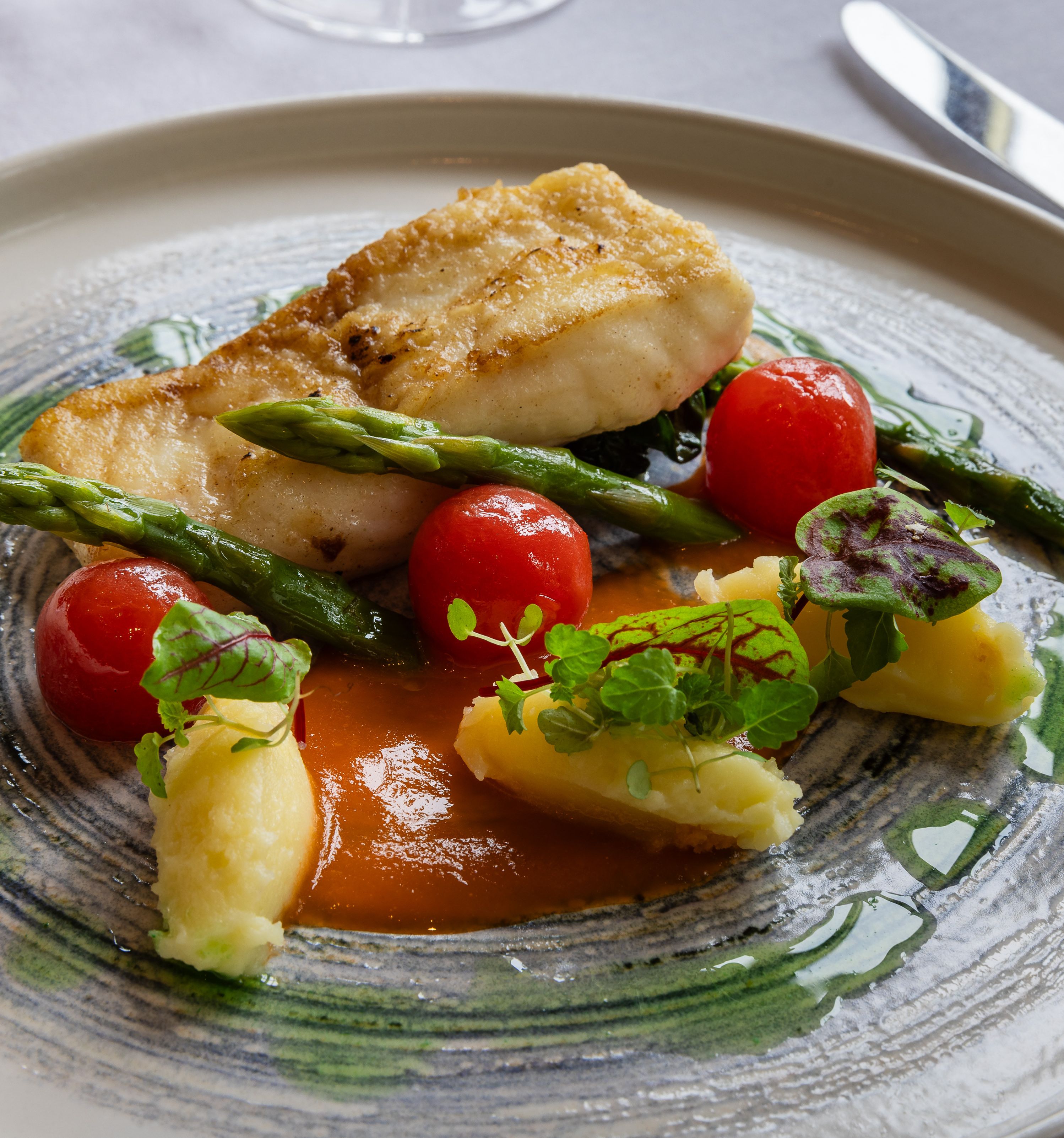 Plated fish fillet with asparagus, cherry tomatoes, mashed potatoes, and sauce, garnished with microgreens.