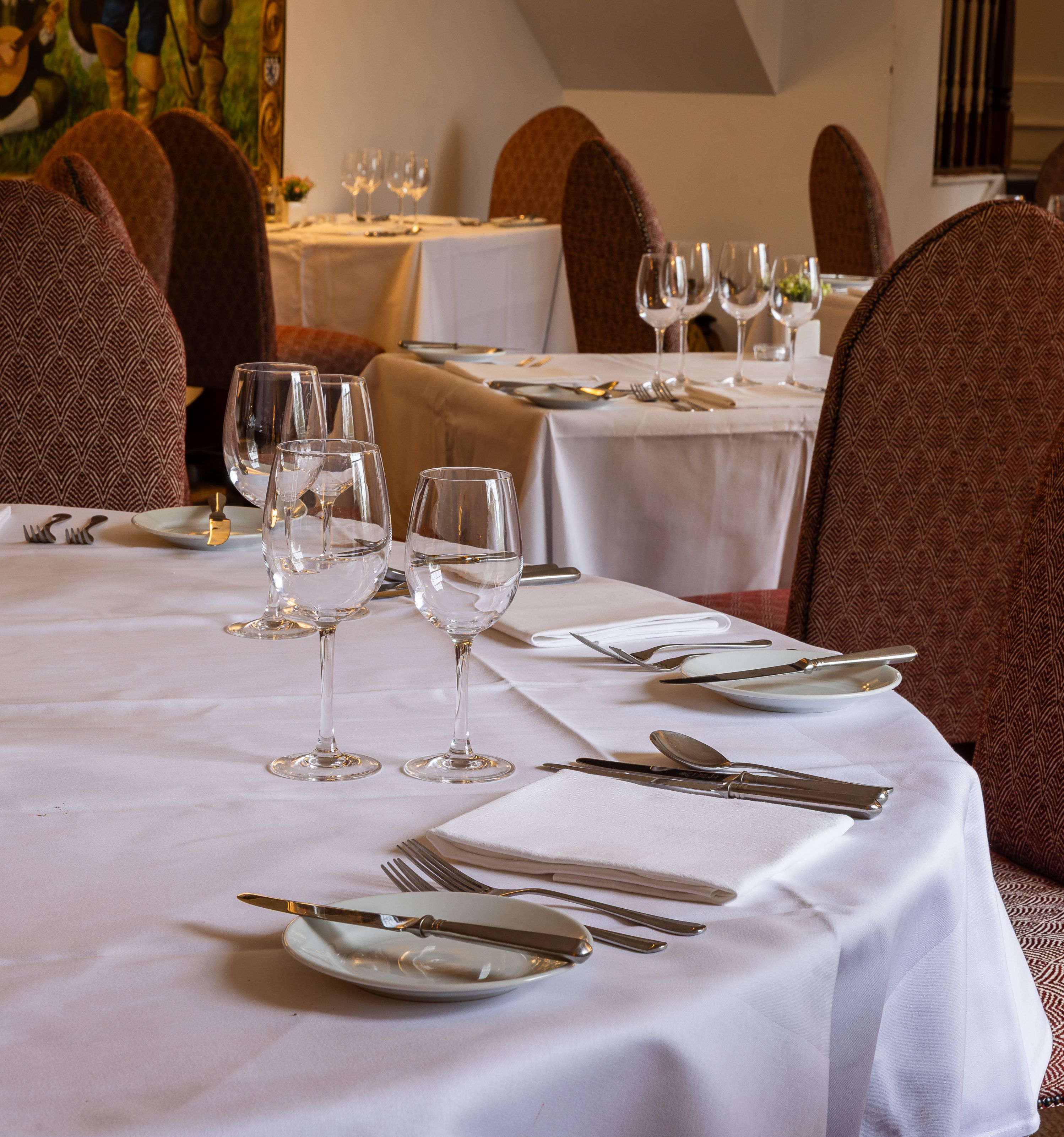 Elegant restaurant interior with tables set for fine dining, featuring wine glasses, white tablecloths, and patterned chairs.