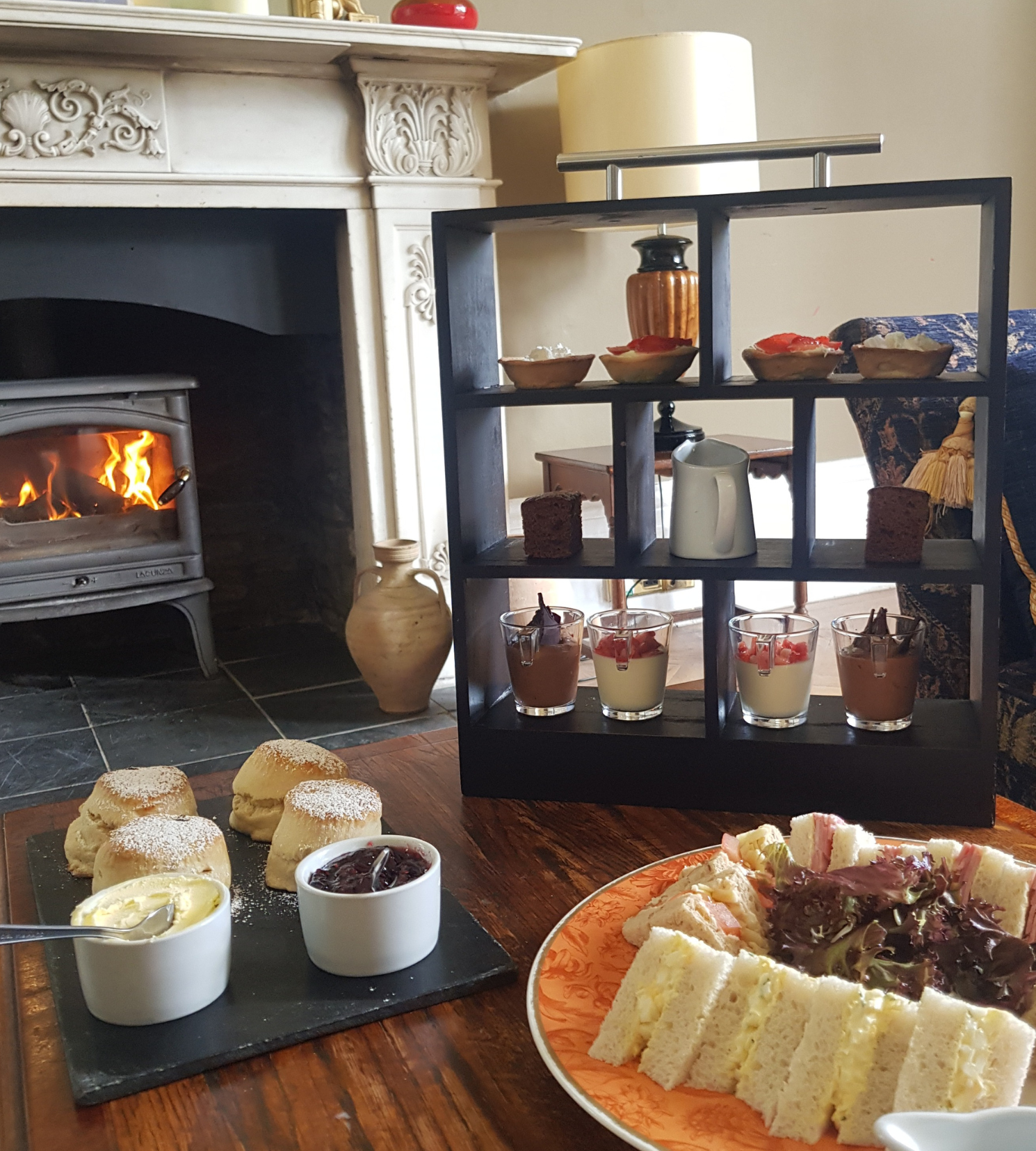 Afternoon tea set with sandwiches, scones, desserts, and a fire in the background