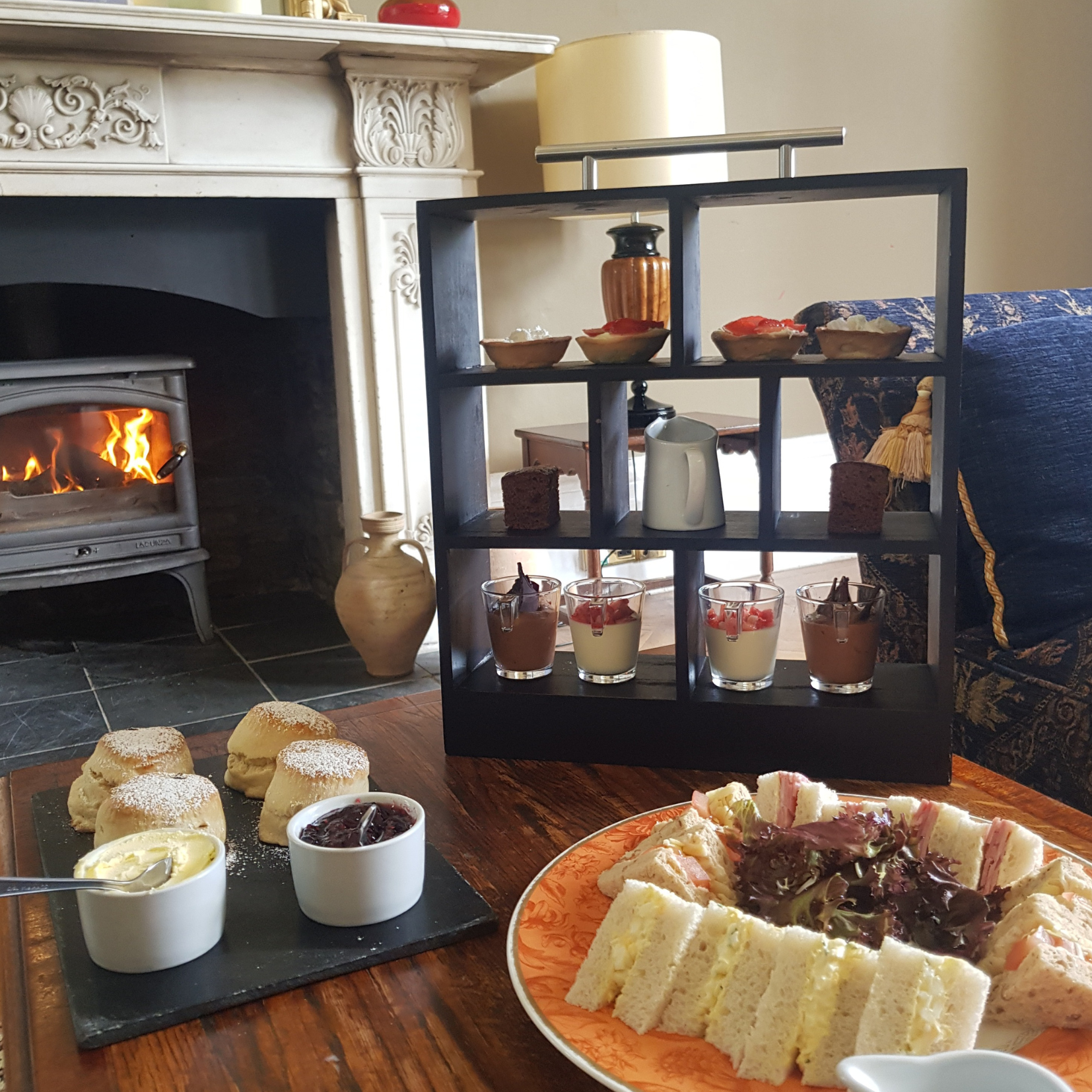 Afternoon tea set with sandwiches, scones, desserts, and a fire in the background