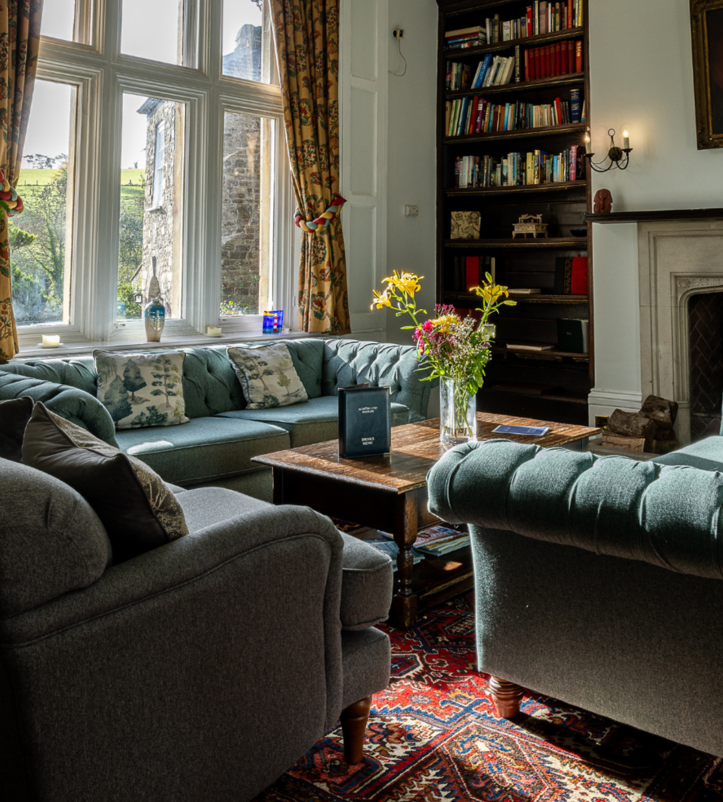 Cozy living room with tufted sofas, wooden table, bookshelves, and a large window with floral curtains.